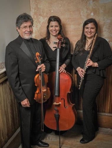 A trio of musicians stands together, holding a violin, cello, and flute, against a textured wall.