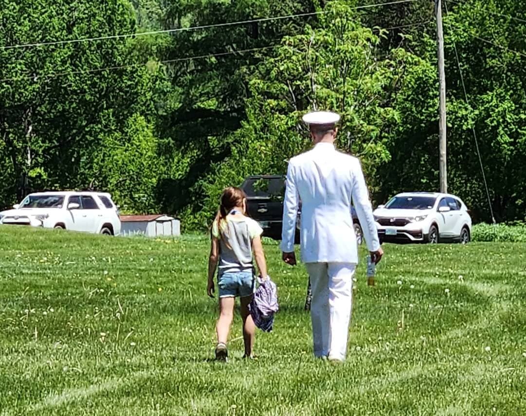 A man in a white military uniform walks alongside a young girl in a grassy field.