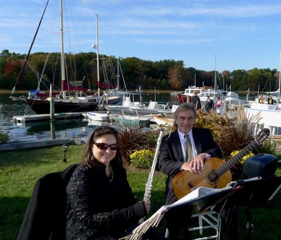 A flutist and guitarist perform by a marina with boats and autumn foliage in the background.
