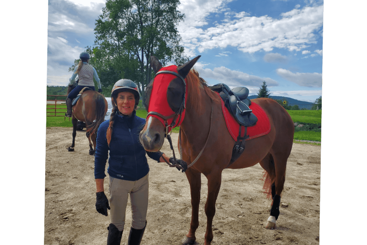 A woman stands beside a brown horse wearing a red saddle and bridle, with another rider in the background.