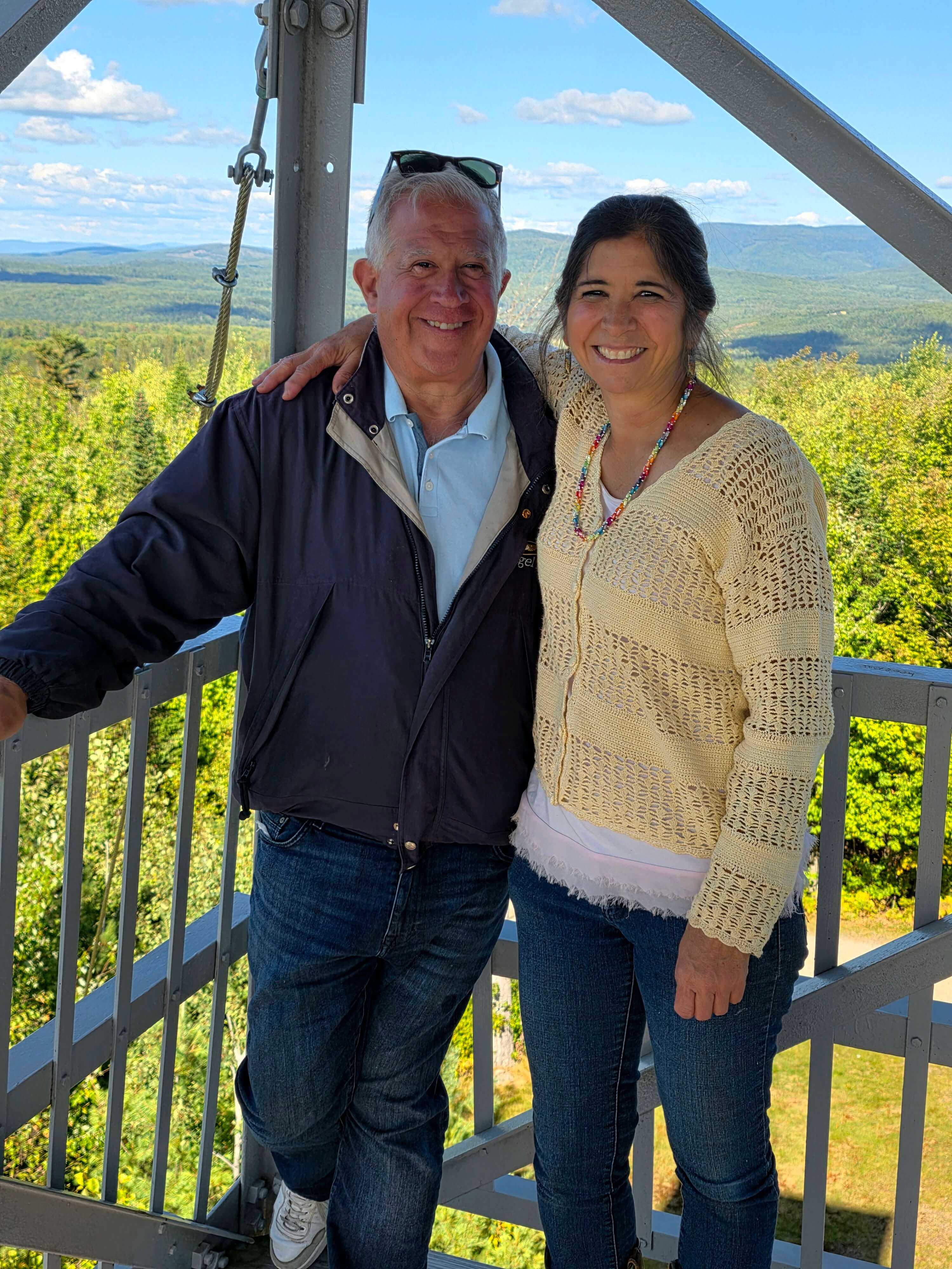 A man and a woman smile together on a viewing platform with a scenic landscape in the background.