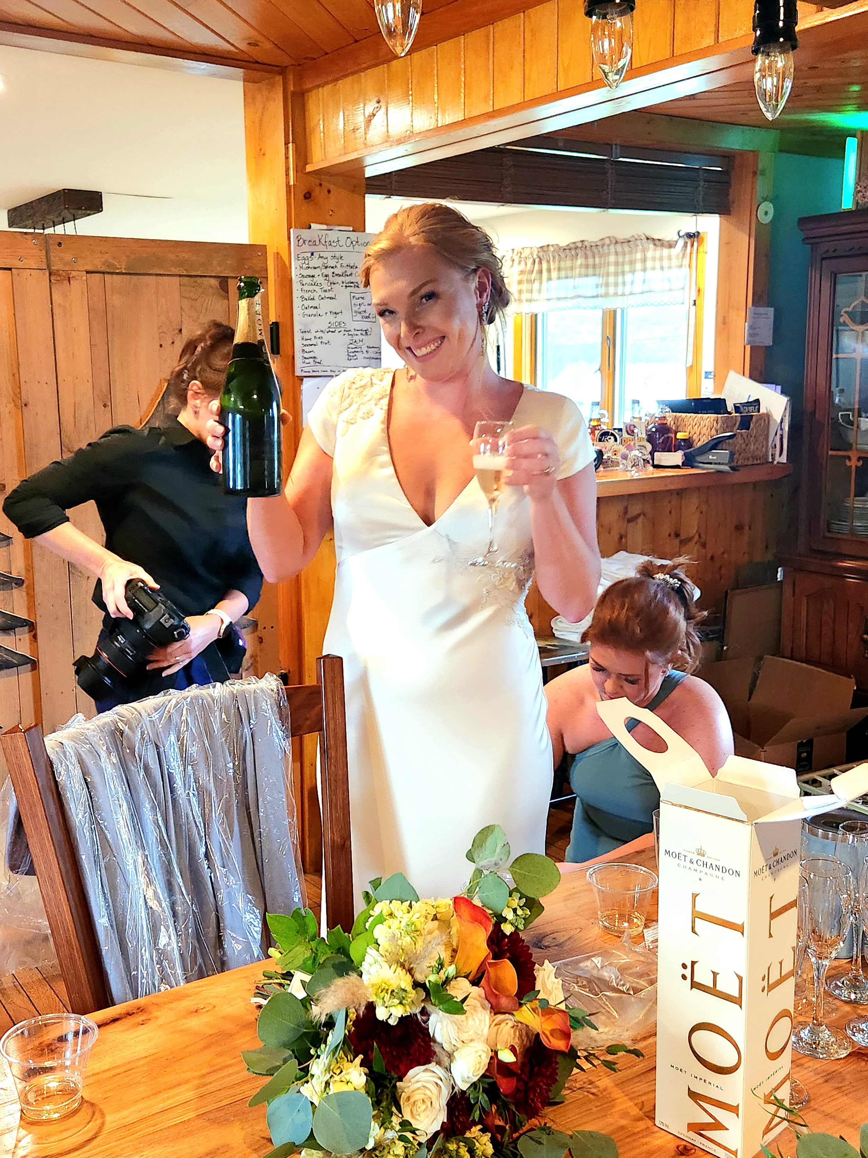 A smiling bride in a white dress raises a champagne glass and a bottle while surrounded by friends and a floral bouquet in a cozy indoor setting.