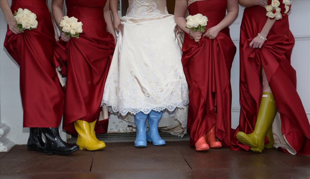 Bridesmaids in red dresses stand together, showcasing colorful rain boots beneath a bride's white gown.
