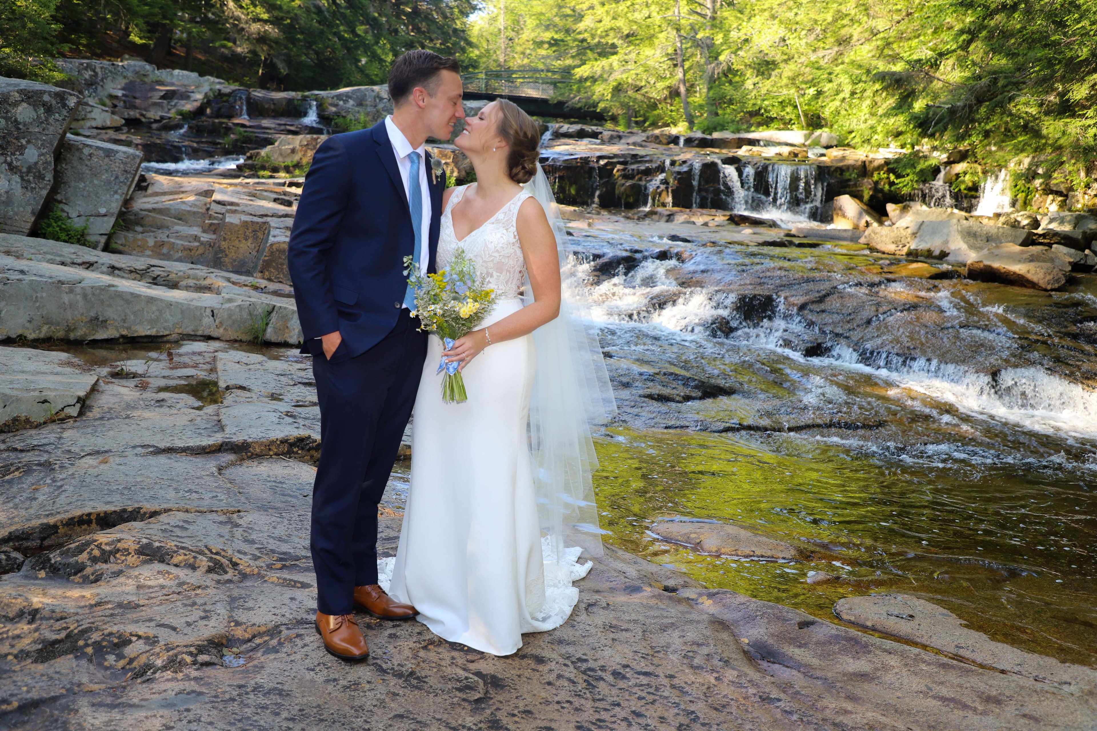A couple shares a romantic kiss by a serene waterfall, surrounded by lush greenery.