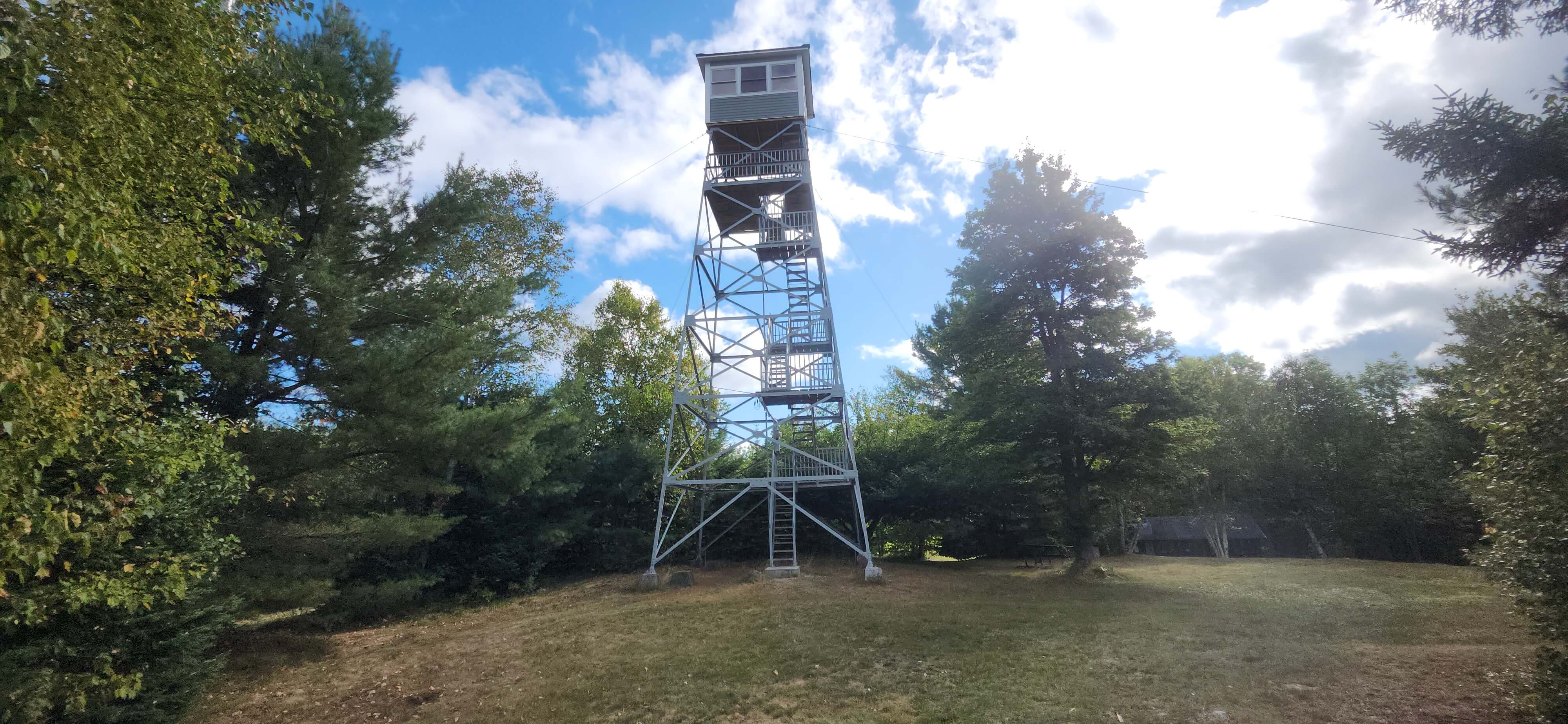 A tall fire lookout tower surrounded by trees against a partly cloudy sky.