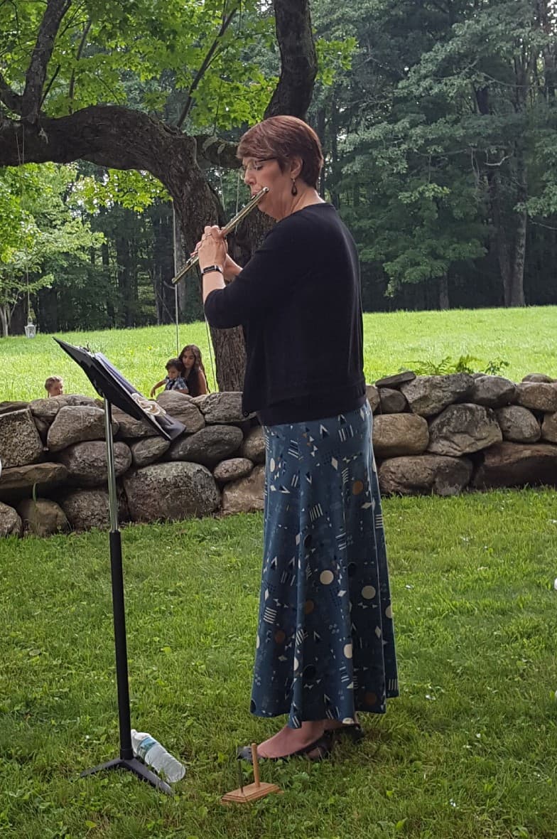 A woman plays the flute outdoors while standing next to a music stand and a stone wall.