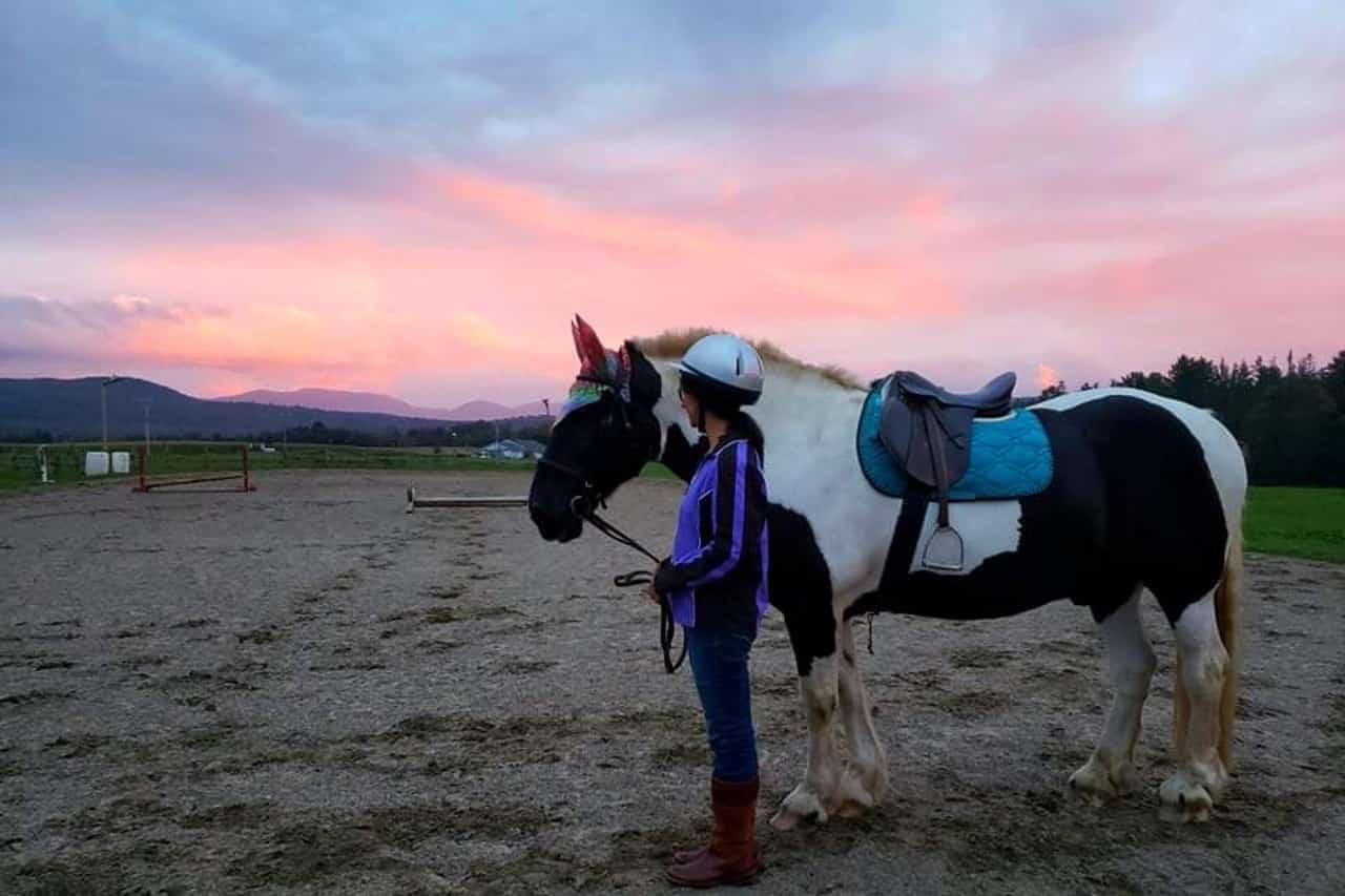 A girl in a helmet stands beside a saddle-covered horse against a colorful sunset.