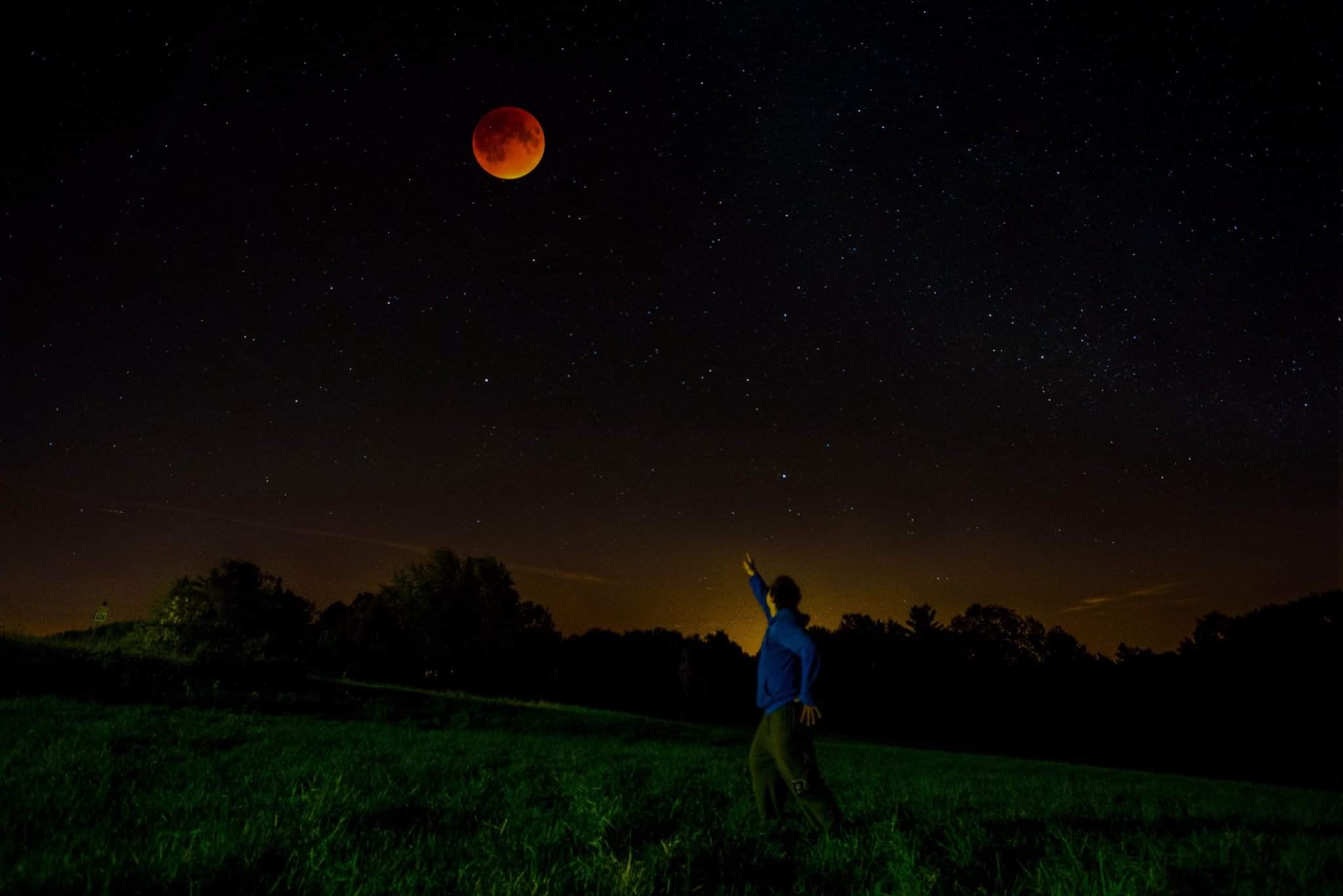 A person stands in a field, pointing at a blood-red moon in a starry night sky.