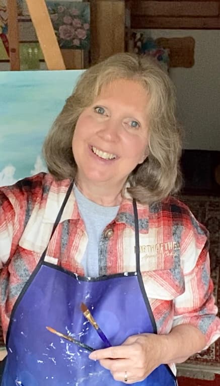 A smiling woman in a plaid shirt and blue apron holds paintbrushes in front of an easel.