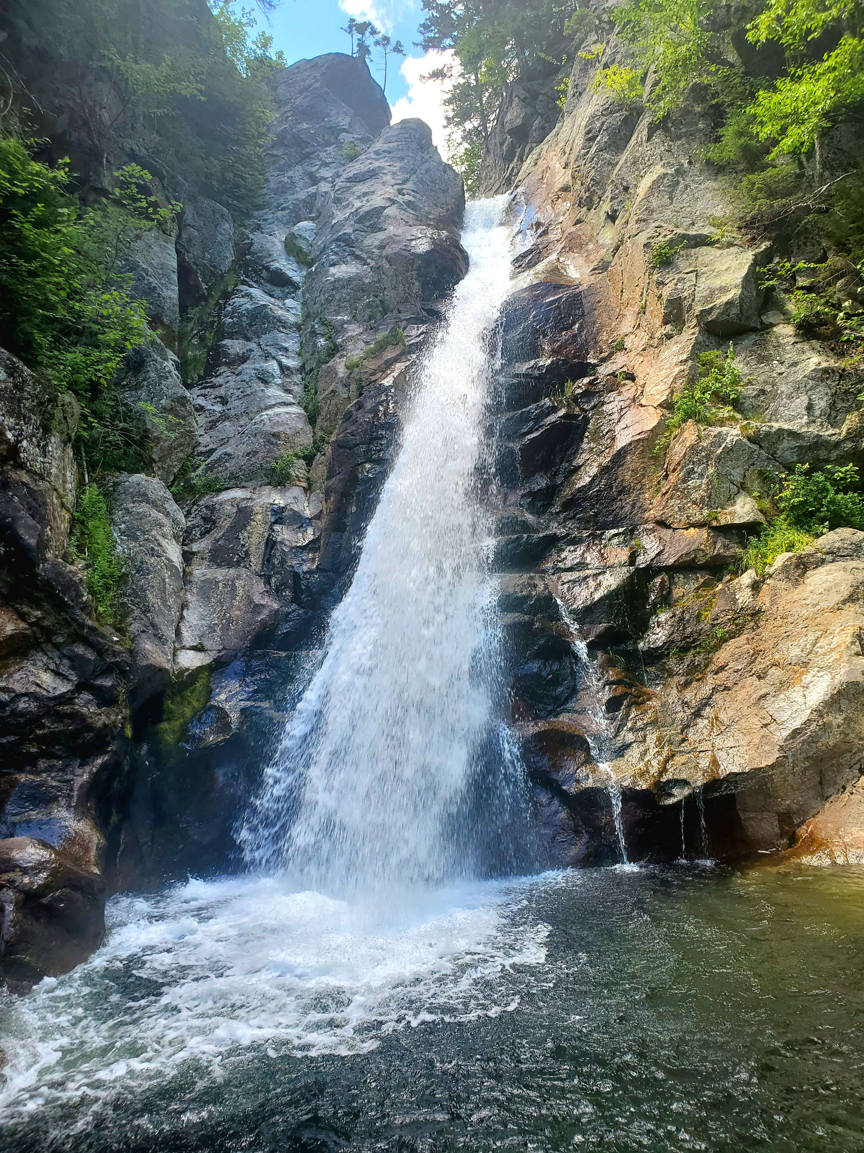 A waterfall cascades over rocky cliffs into a serene pool surrounded by lush greenery.