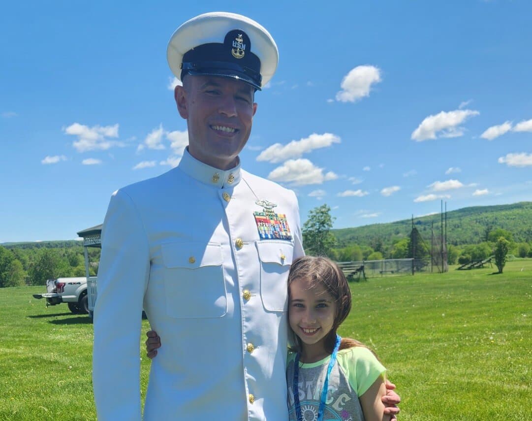 A smiling military officer in uniform poses with a young girl on a sunny day.