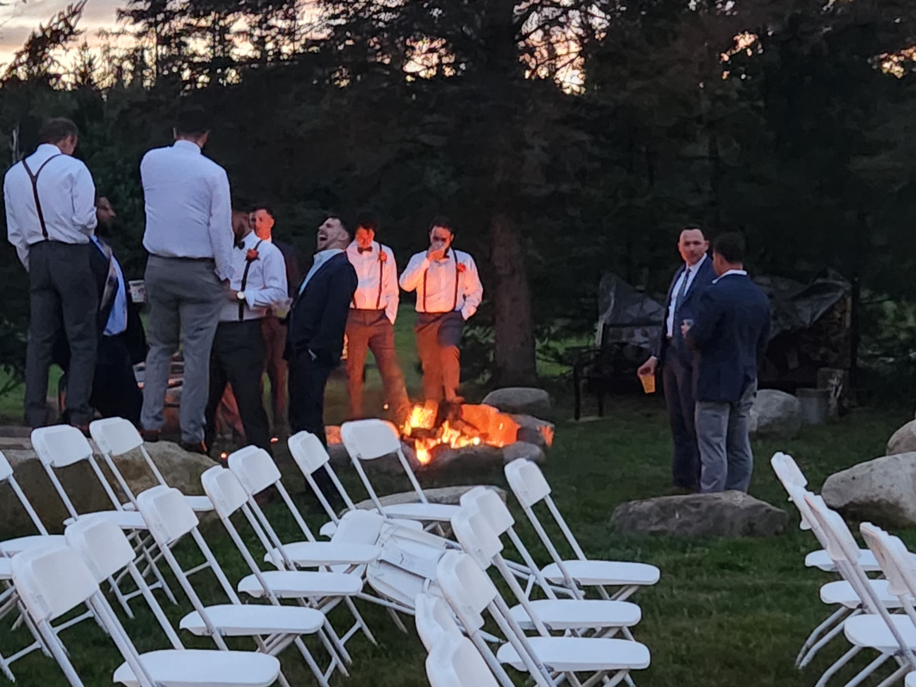 A group of men in formal attire gather around a fire in an outdoor setting with empty chairs nearby.