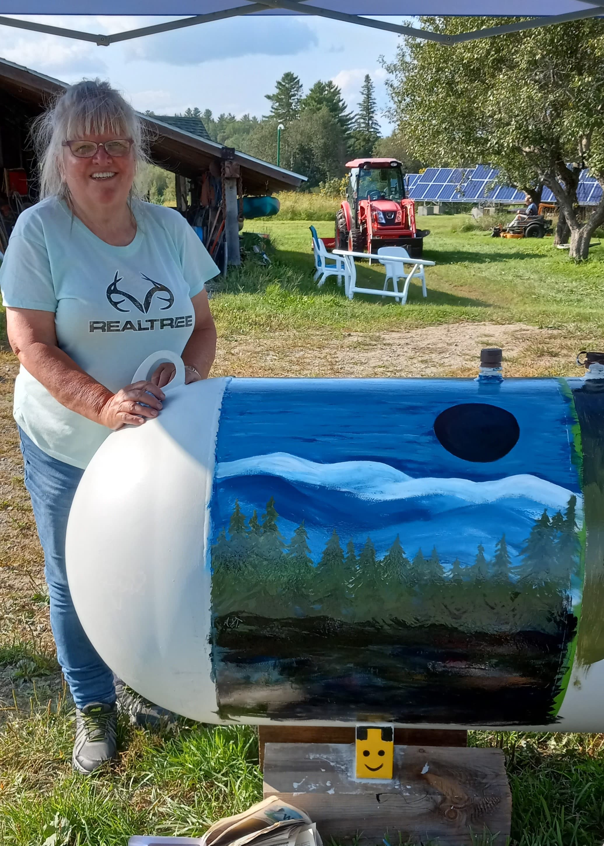 A woman stands next to a painted propane tank showcasing a landscape scene, with a tractor and solar panels in the background.