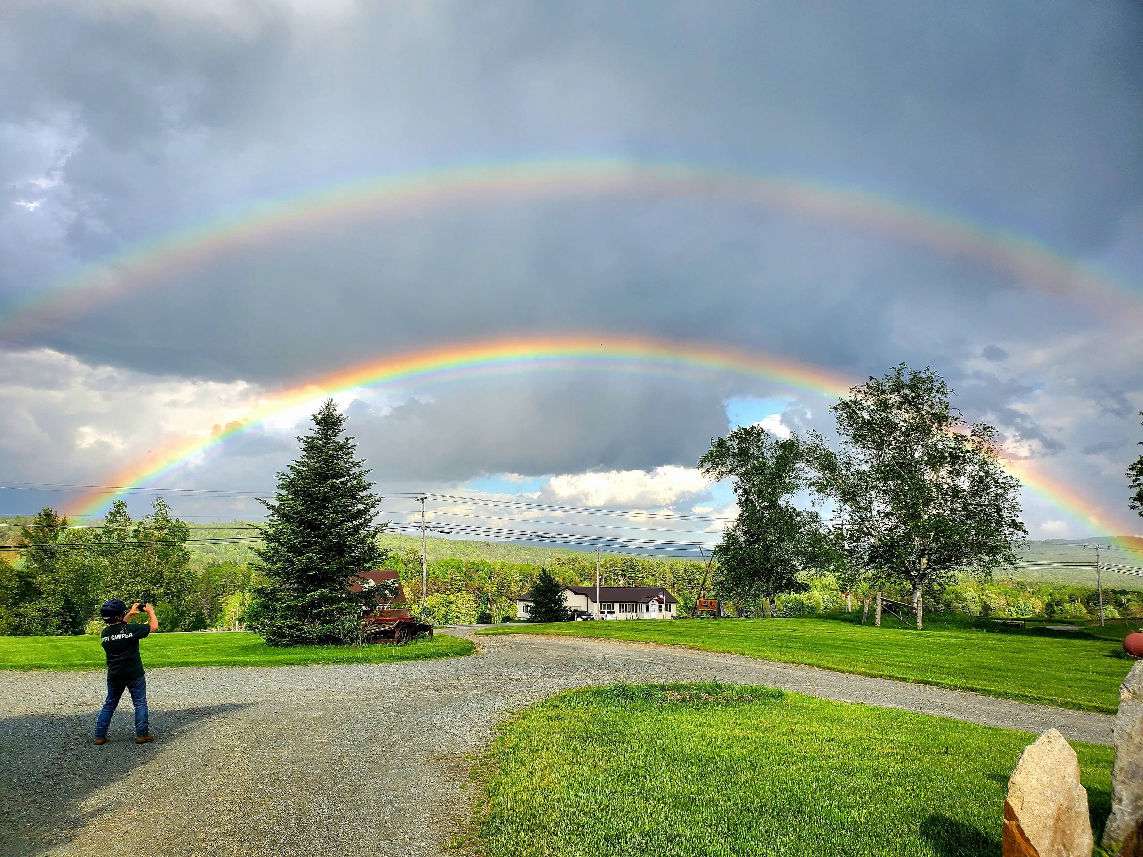 A person stands on a gravel path admiring a double rainbow over a green landscape and distant hills.