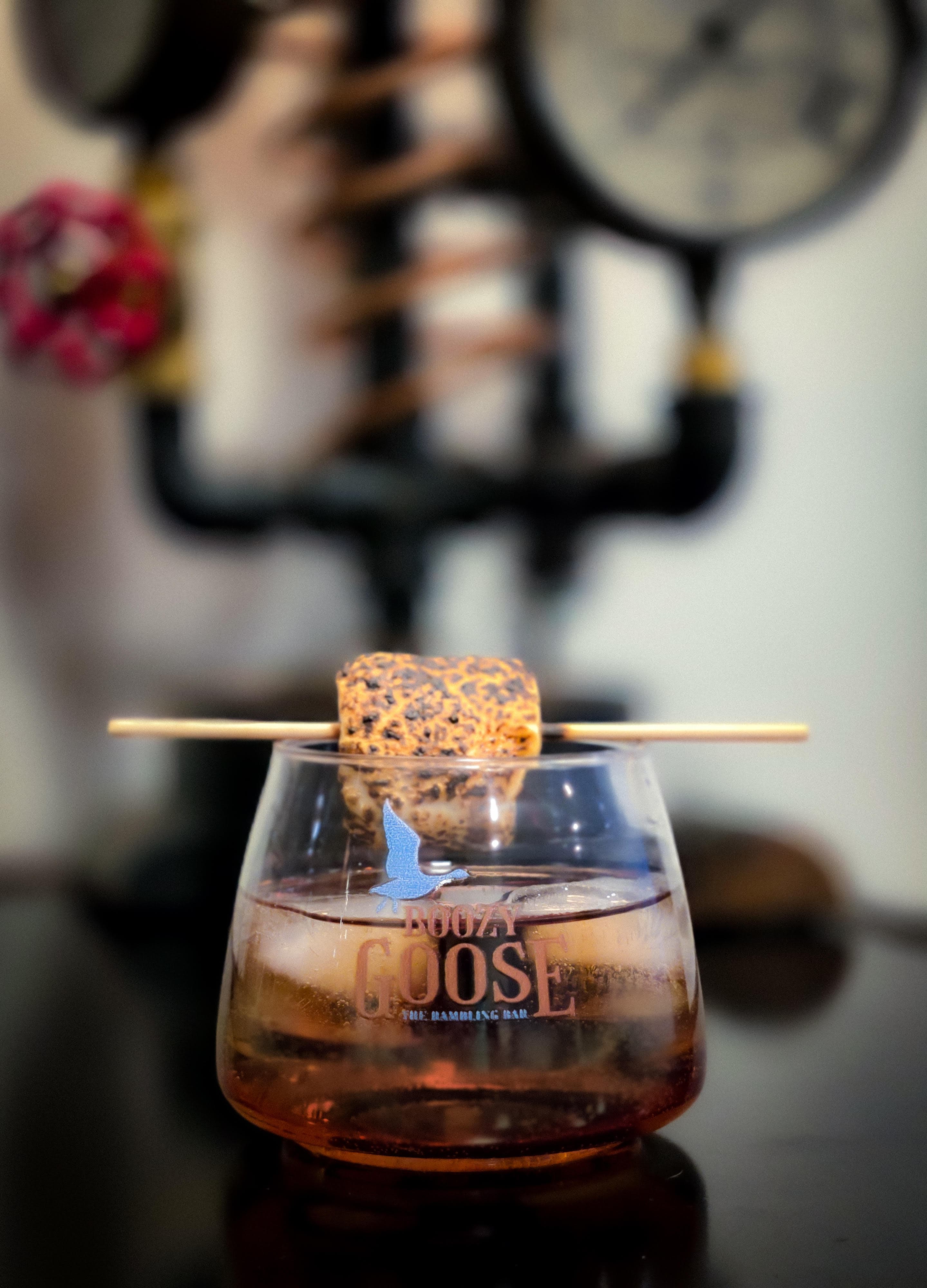 A cocktail with a toasted marshmallow on a skewer rests in a glass branded with "Boozy Goose."