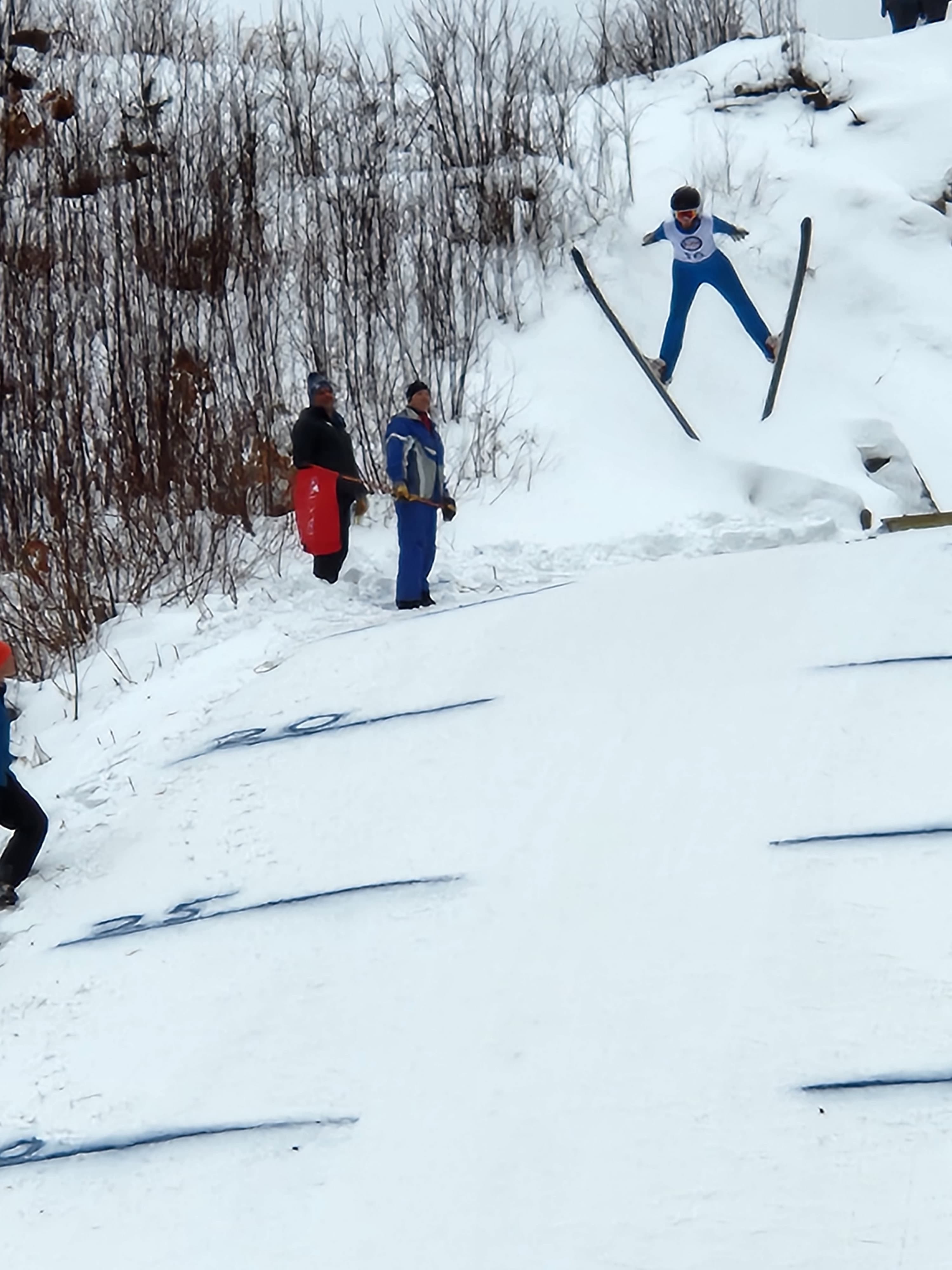 A skier in mid-air jumps off a snowy ramp as onlookers observe.