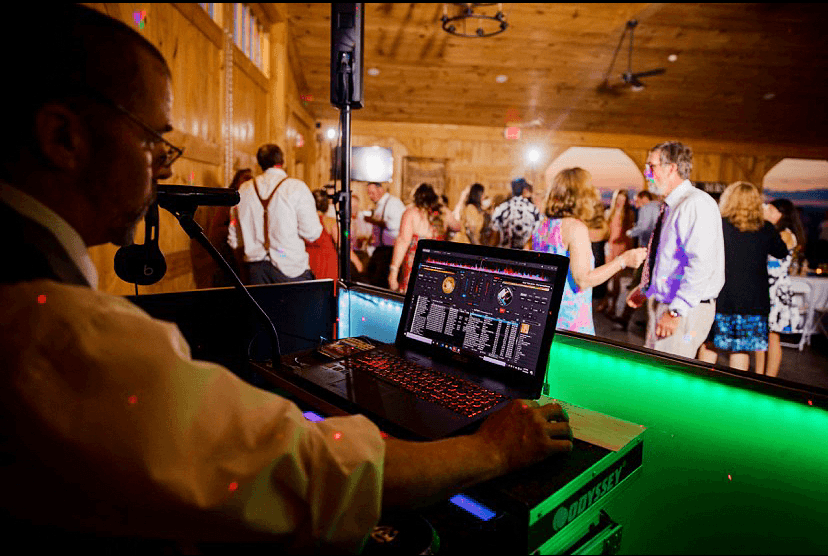 A DJ mixes music at a lively event with guests dancing in the background.