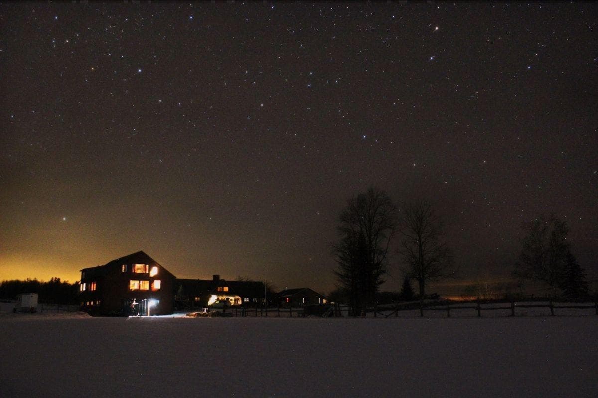 A snowy landscape at night featuring a house illuminated by lights against a starry sky.
