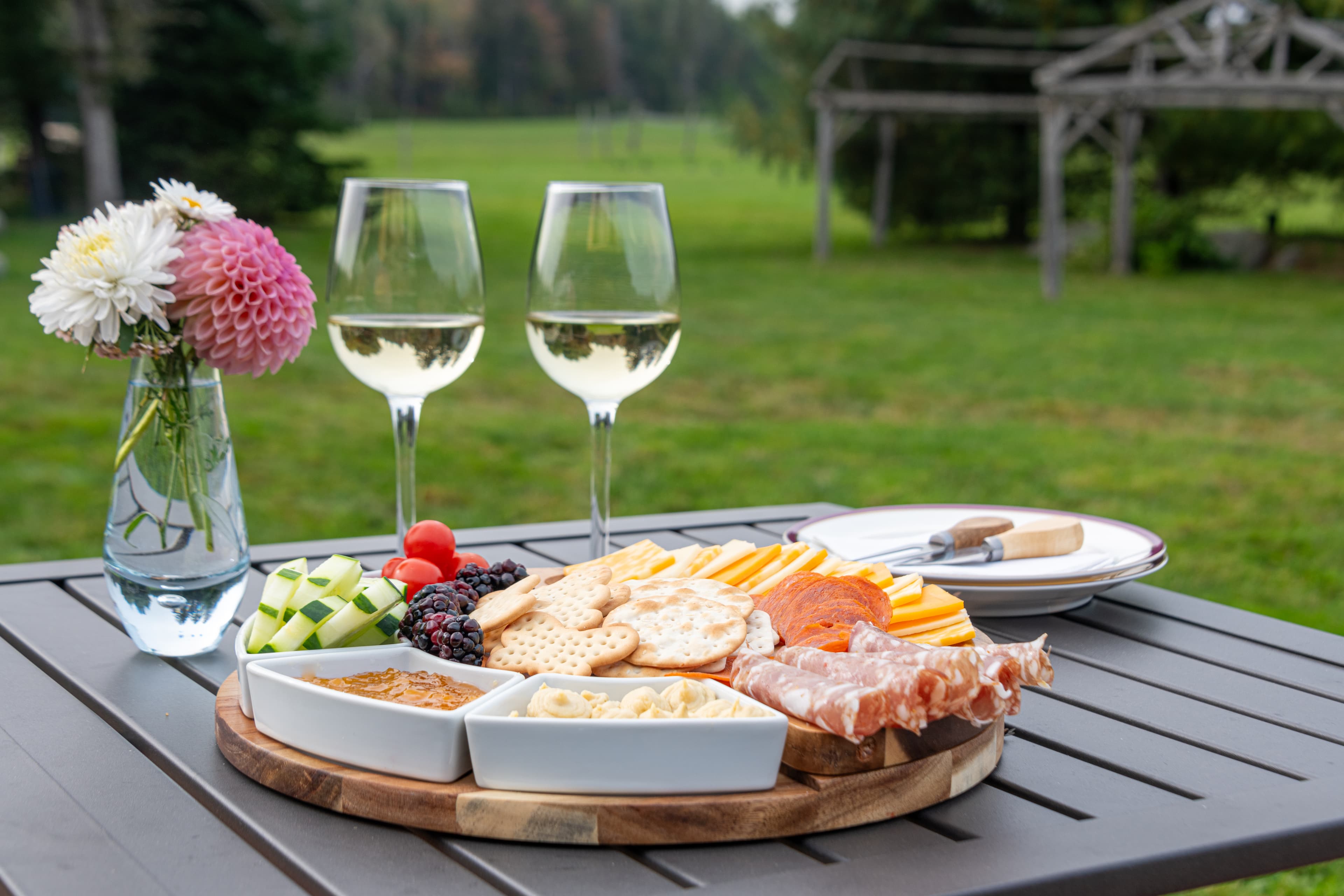 Round wood platter filled with charcuterie items including meats, cheeses, vegetables and fruit, with two glasses of wine.