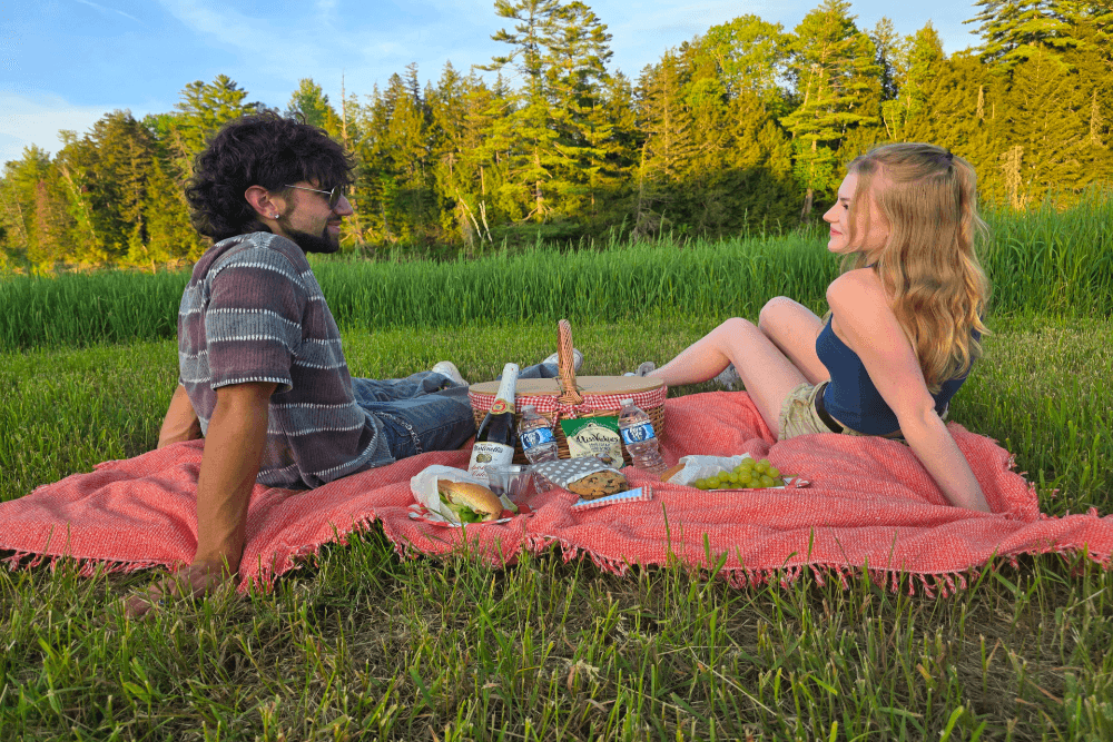 A couple enjoys a picnic on a blanket in a grassy field surrounded by trees.