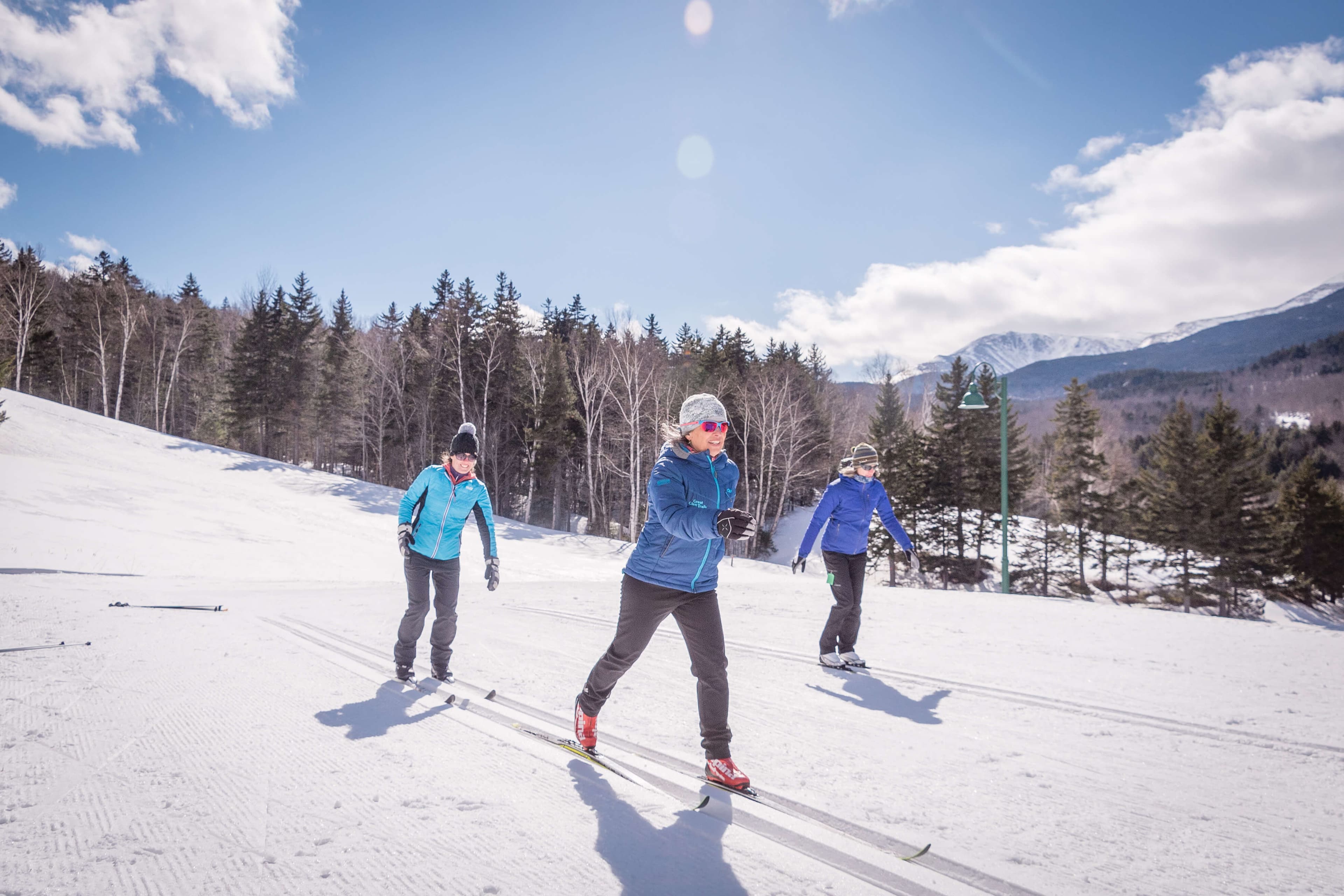 Three people cross-country skiing on a snowy landscape surrounded by trees and mountains. Three people cross-country skiing on a snowy landscape surrounded by trees and mountains.