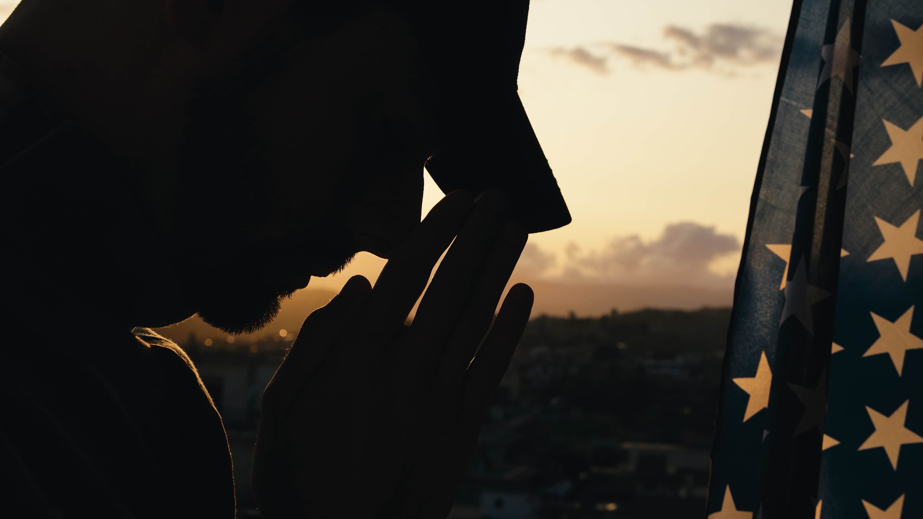 A silhouette of a man in a cap praying in front of an American flag during sunset.