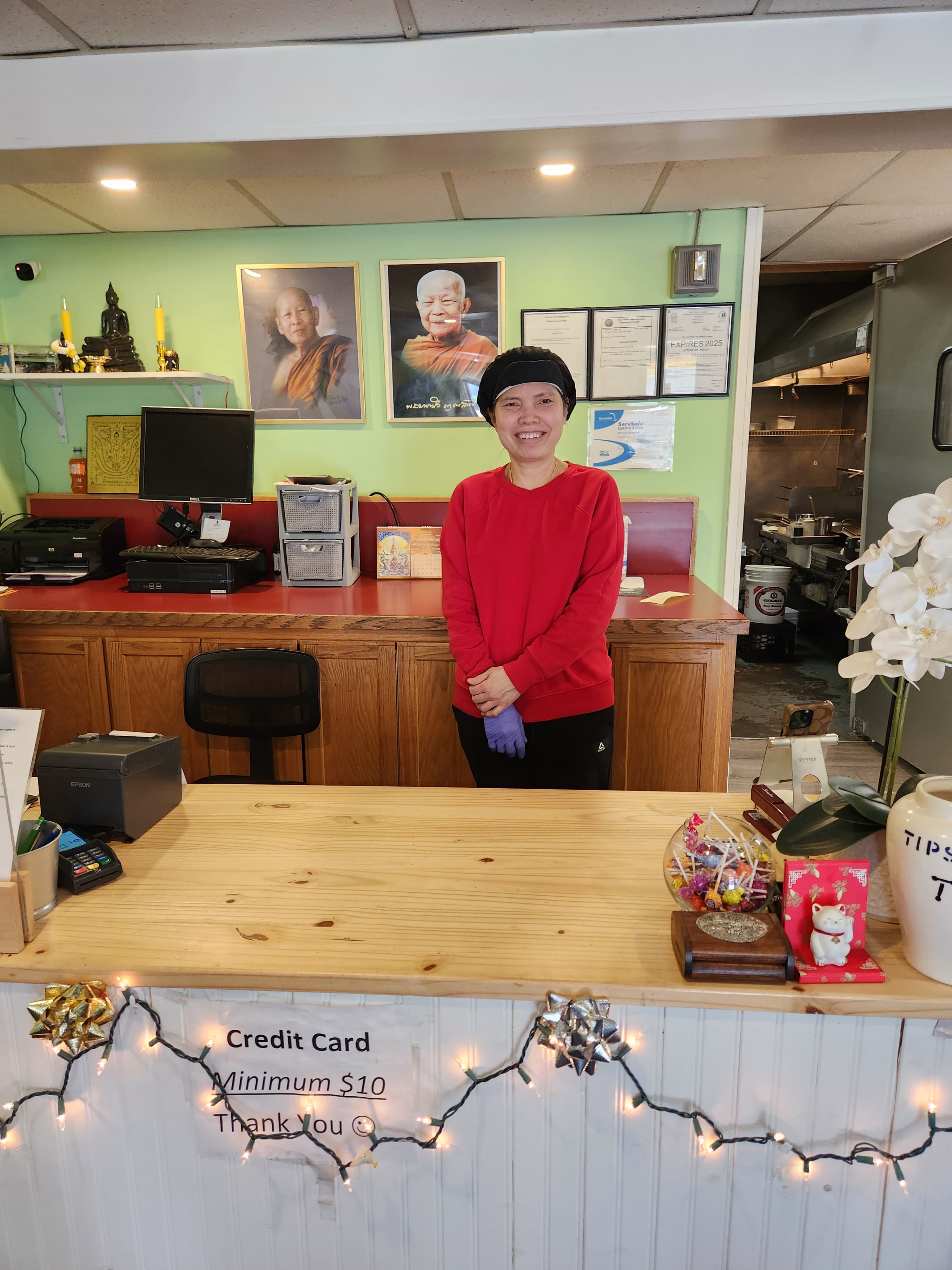 A smiling person in a red sweater stands behind a wooden counter in a restaurant, with decor and kitchen space visible in the background.