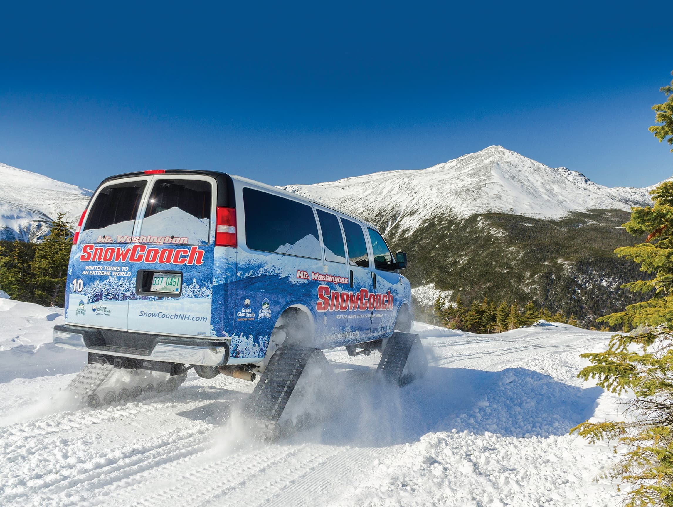 A SnowCoach van traverses a snowy mountain trail with a clear blue sky in the background.