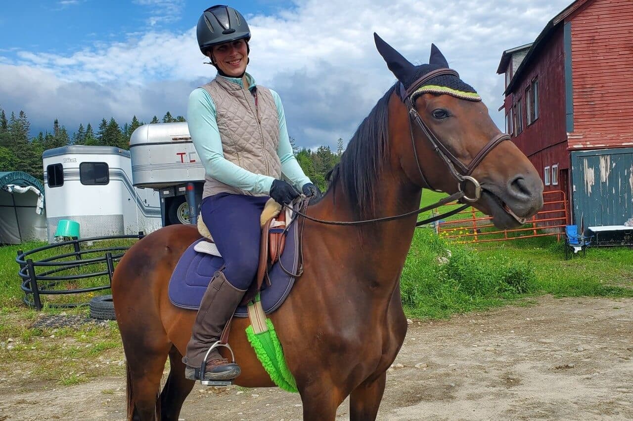 A woman in riding gear smiles while seated on a brown horse in an outdoor setting.