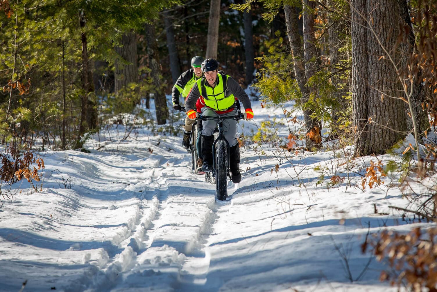 Two cyclists in bright gear ride on a snowy forest trail.