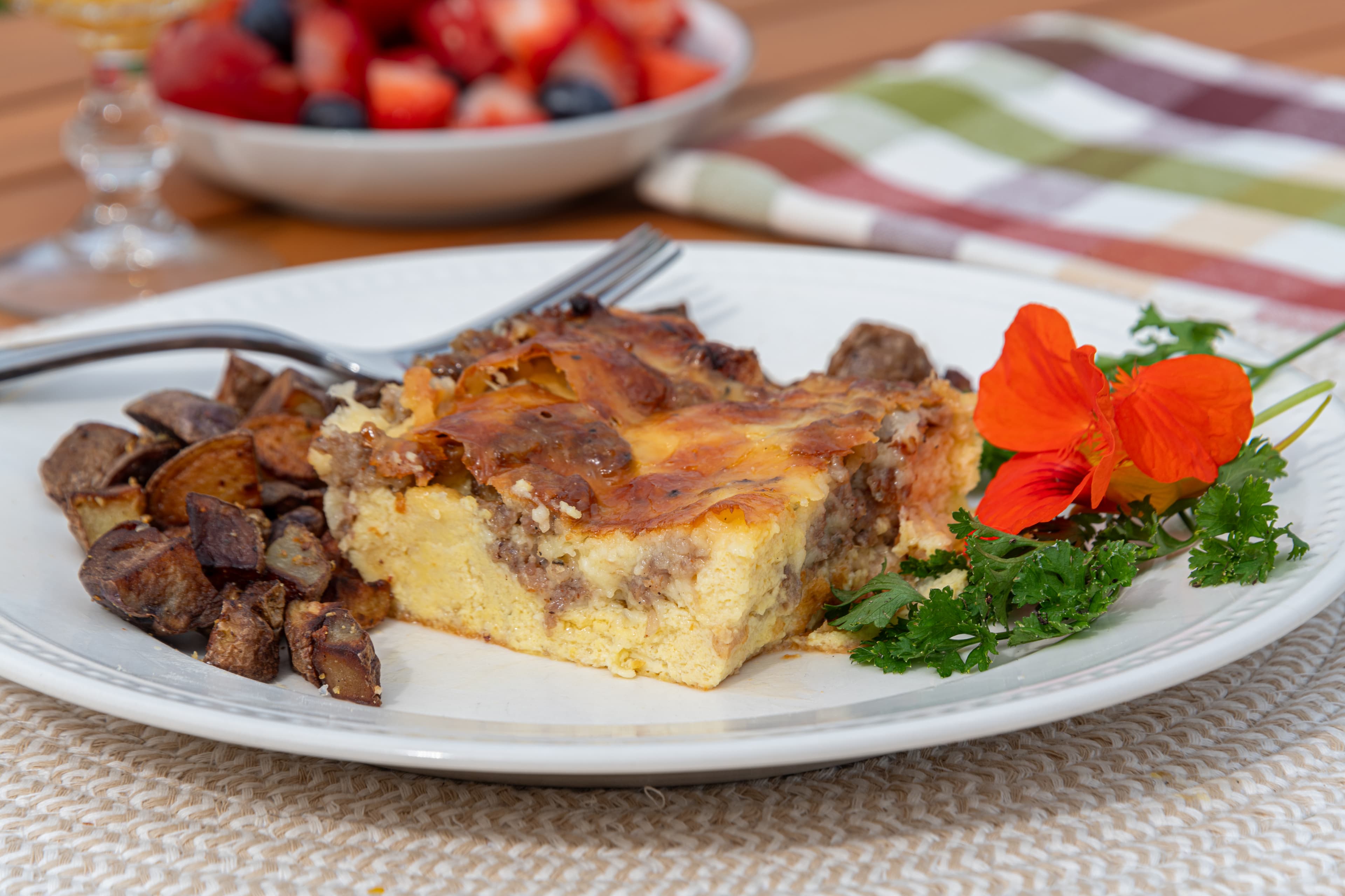 A square of a golden brown egg and sausage casserole with diced potatoes and a bowl of berries.