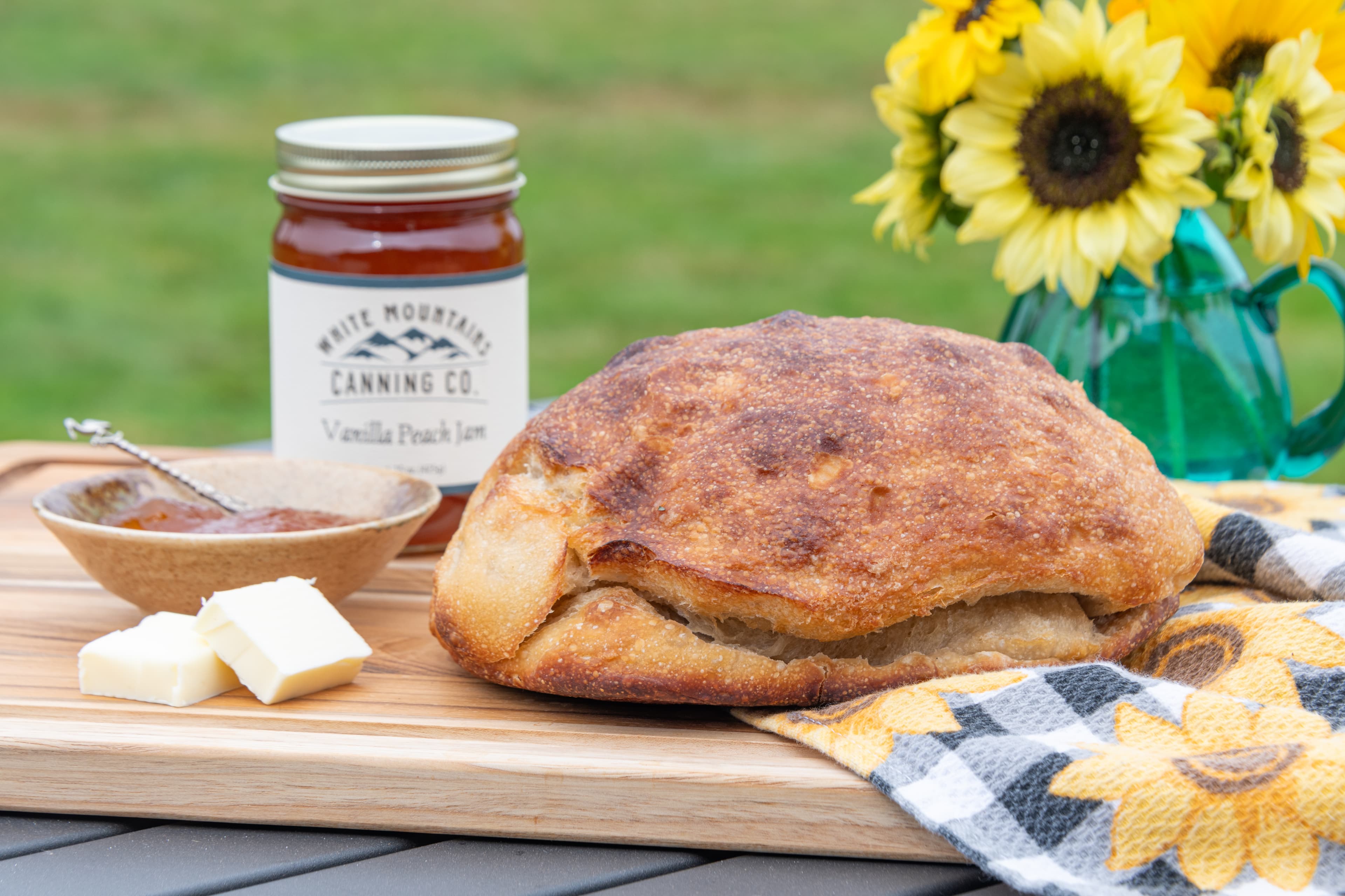Golden brown, freshly baked loaf of sourdough bread on a cutting board with a jar of jam next to a vase of yellow sunflowers.
