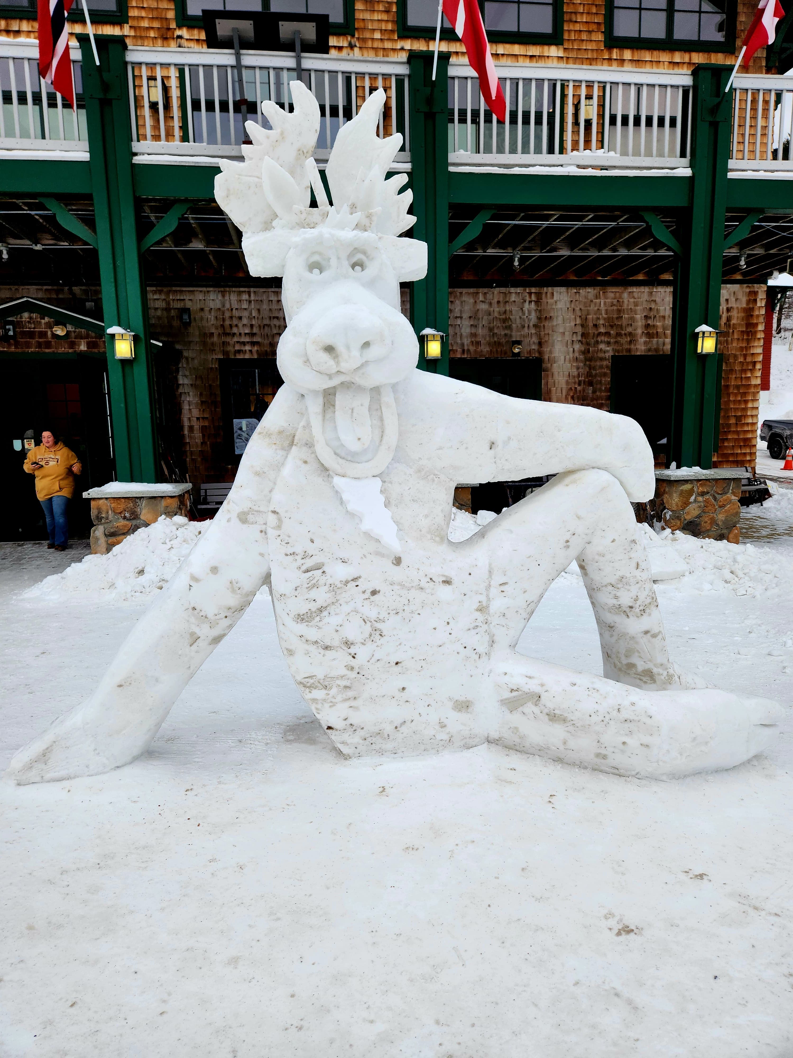 A large, whimsical snow sculpture of a moose sitting down in front of a building.