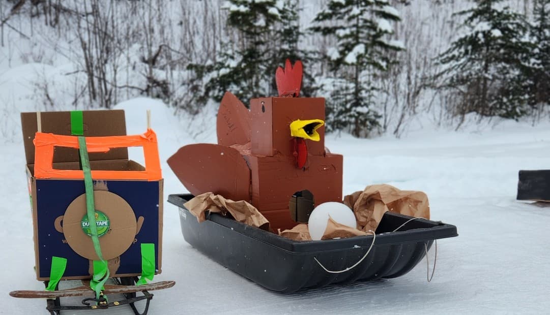 A cardboard turkey and a colorful sled sit on snow-covered ground surrounded by pine trees.