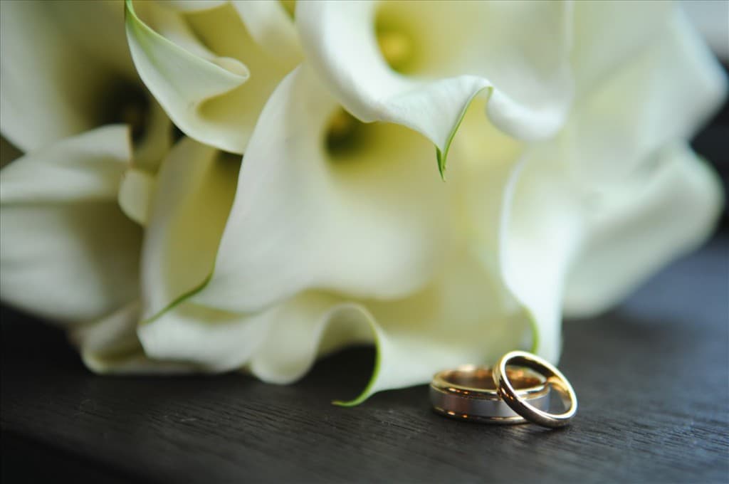 A close-up of two wedding rings resting on a dark surface beside white calla lilies. A close-up of two wedding rings resting on a dark surface beside white calla lilies.