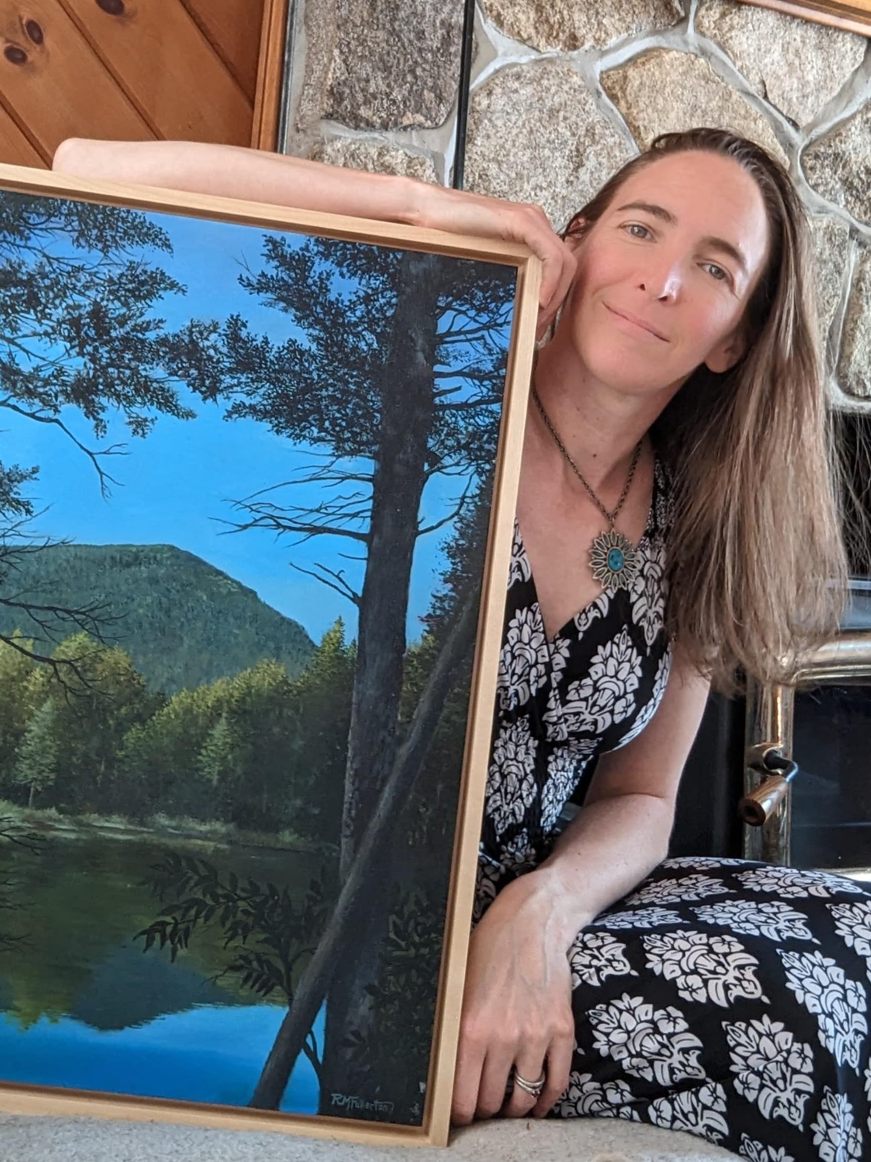 A woman in a patterned dress holds up a framed landscape painting in front of a stone fireplace.