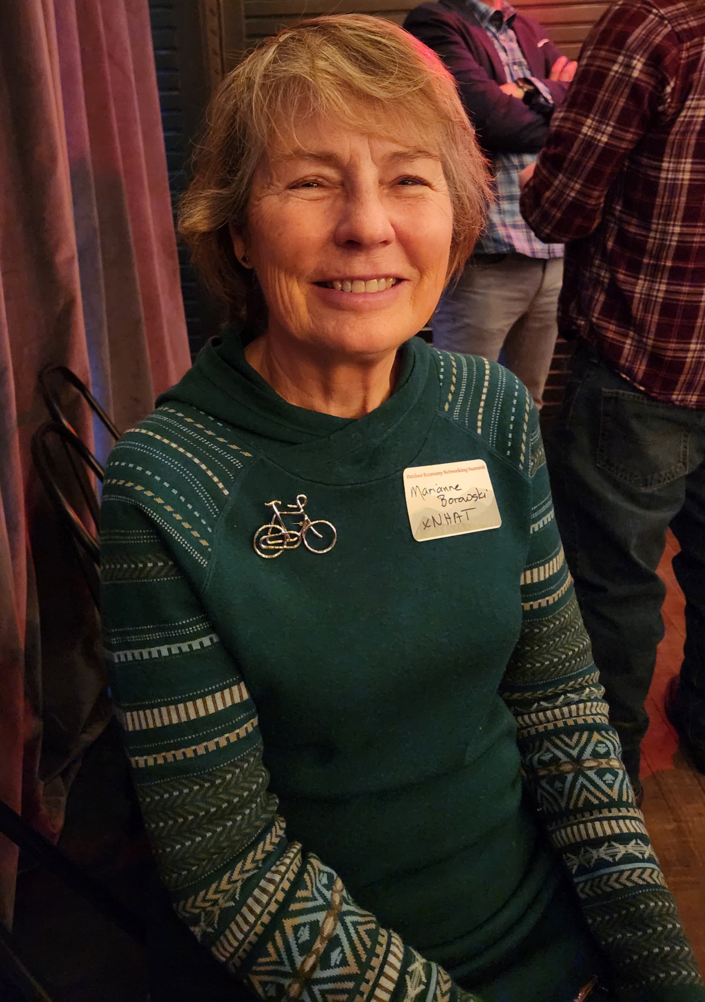 A smiling woman with short hair wearing a green sweater adorned with a bicycle pin.