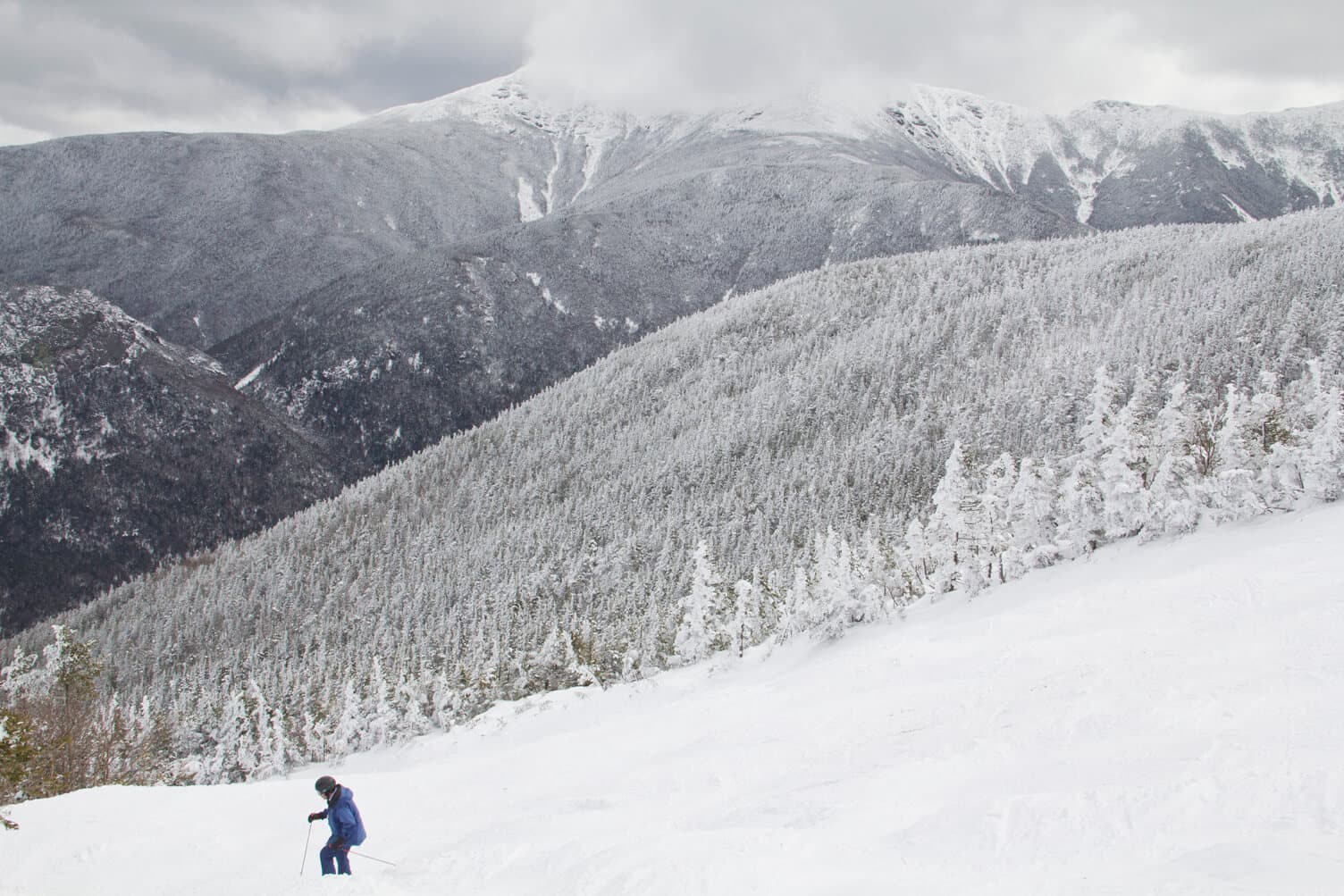 A skier descends a snowy slope with mountains in the background.
