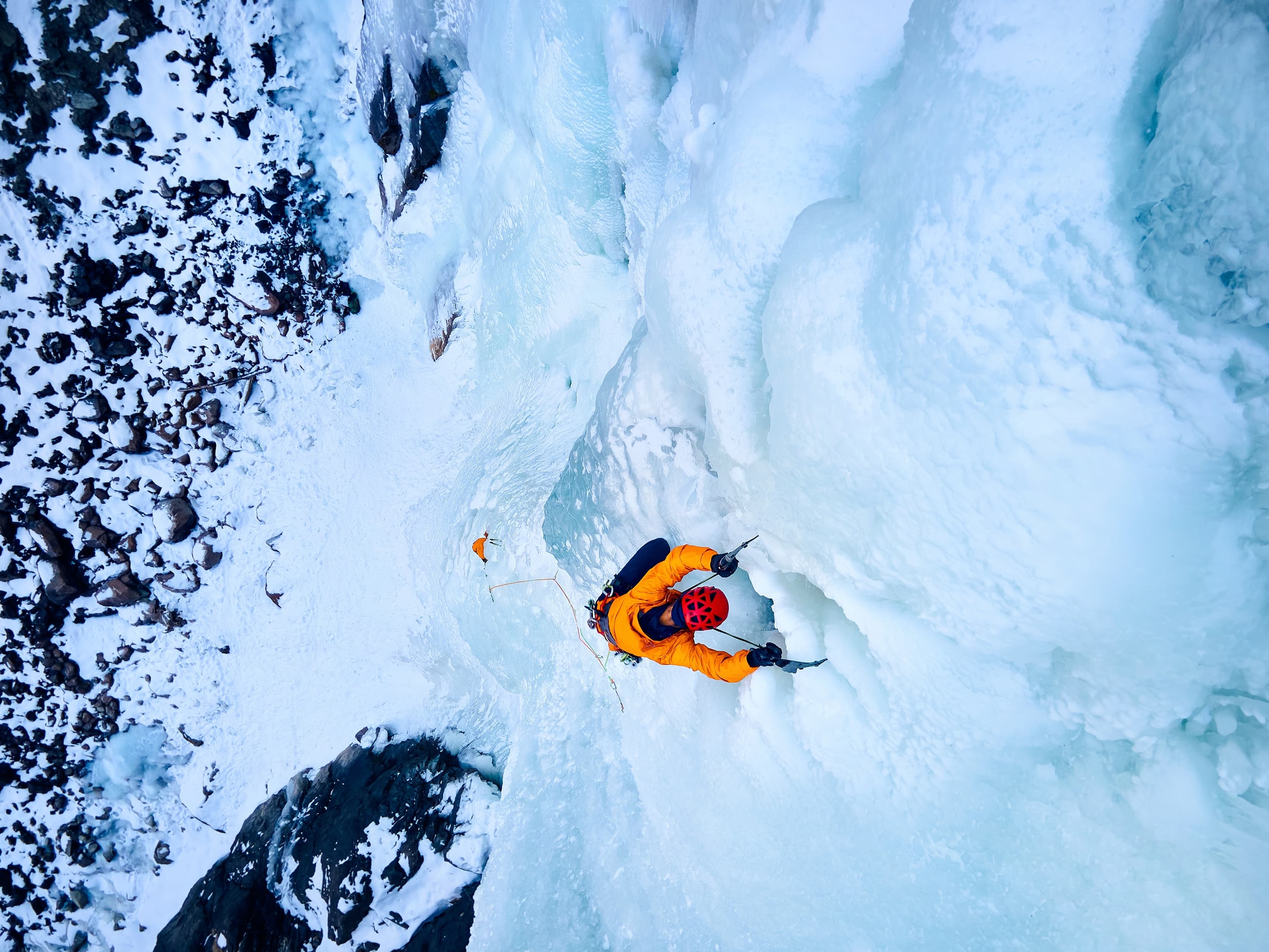 A climber in an orange jacket ascends an icy wall surrounded by snow and rocky terrain.