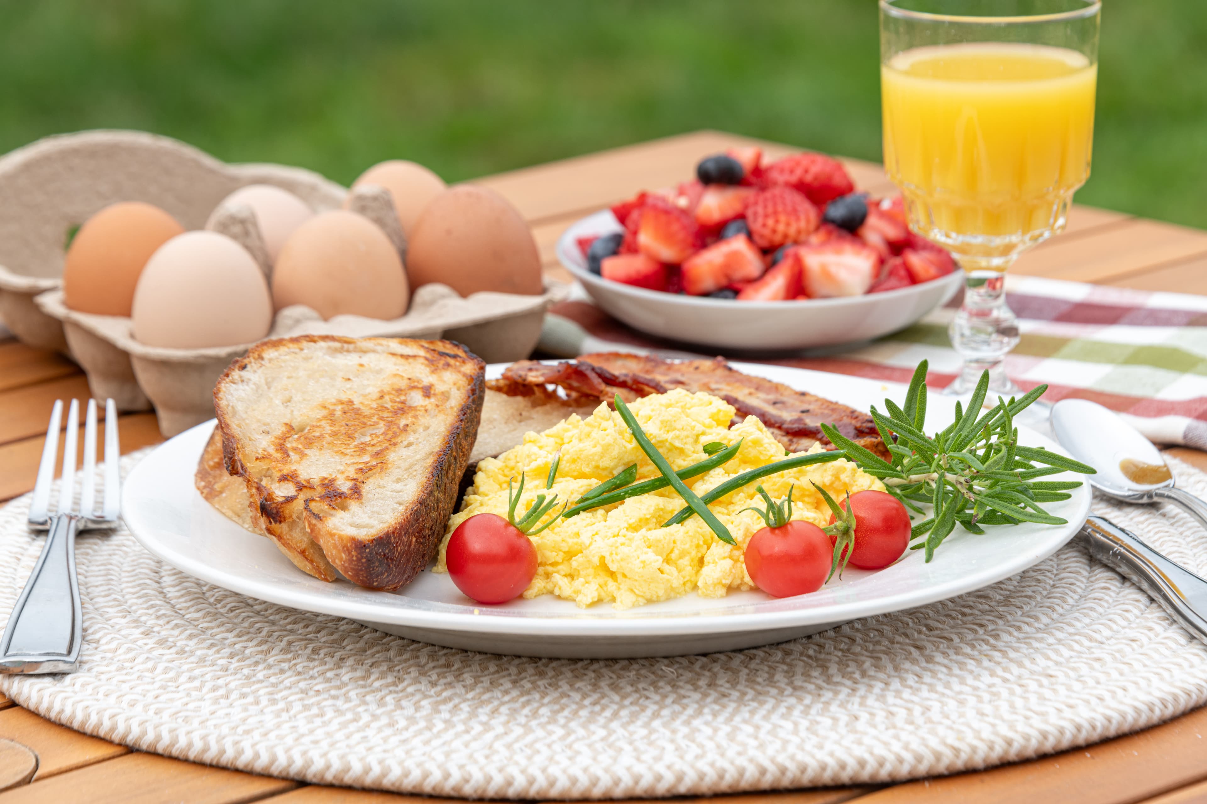 Breakfast plate of scrambled eggs. toast, bacon, cherry tomatoes, bowl of berries and a glass of orange juice.