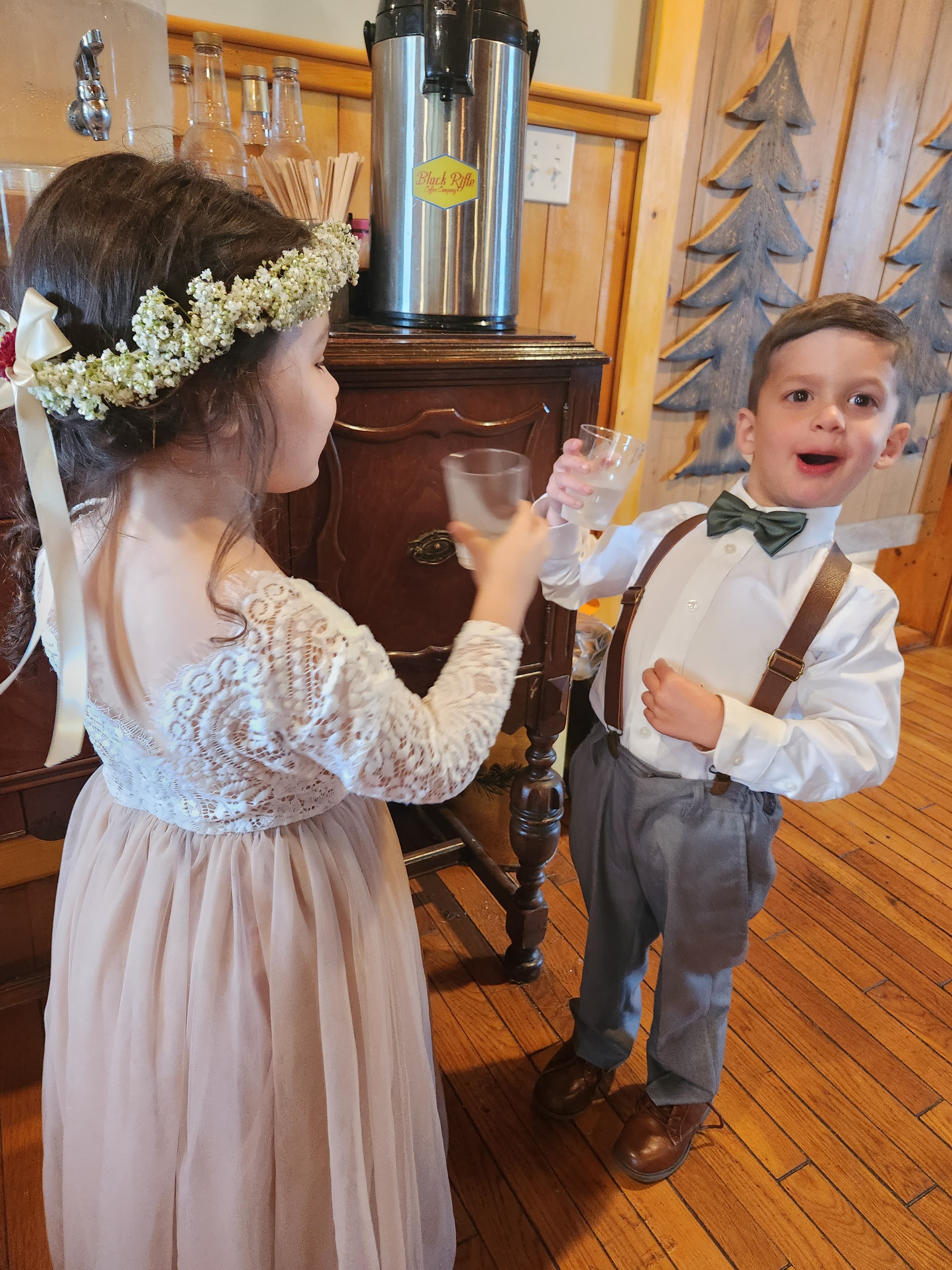 Two children, a girl in a floral crown and a boy in suspenders, are toasting with glasses at a festive gathering.