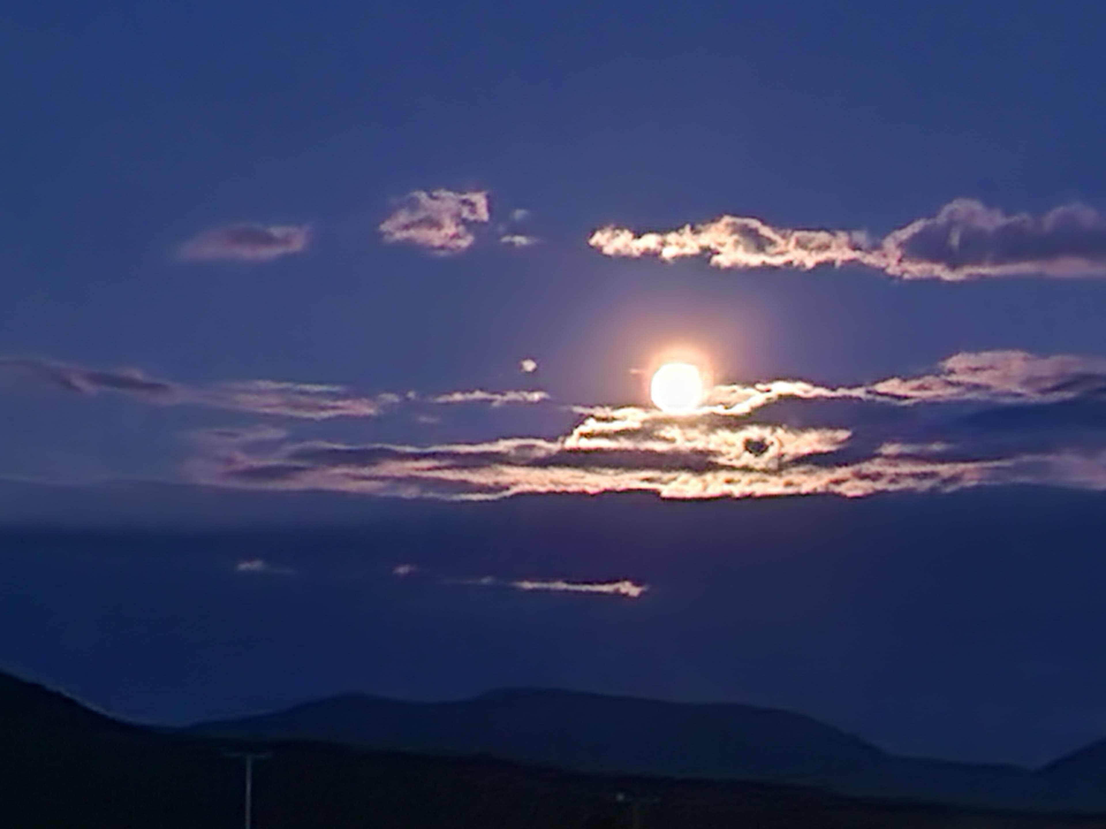A full moon shines brightly among wispy clouds in a twilight sky.