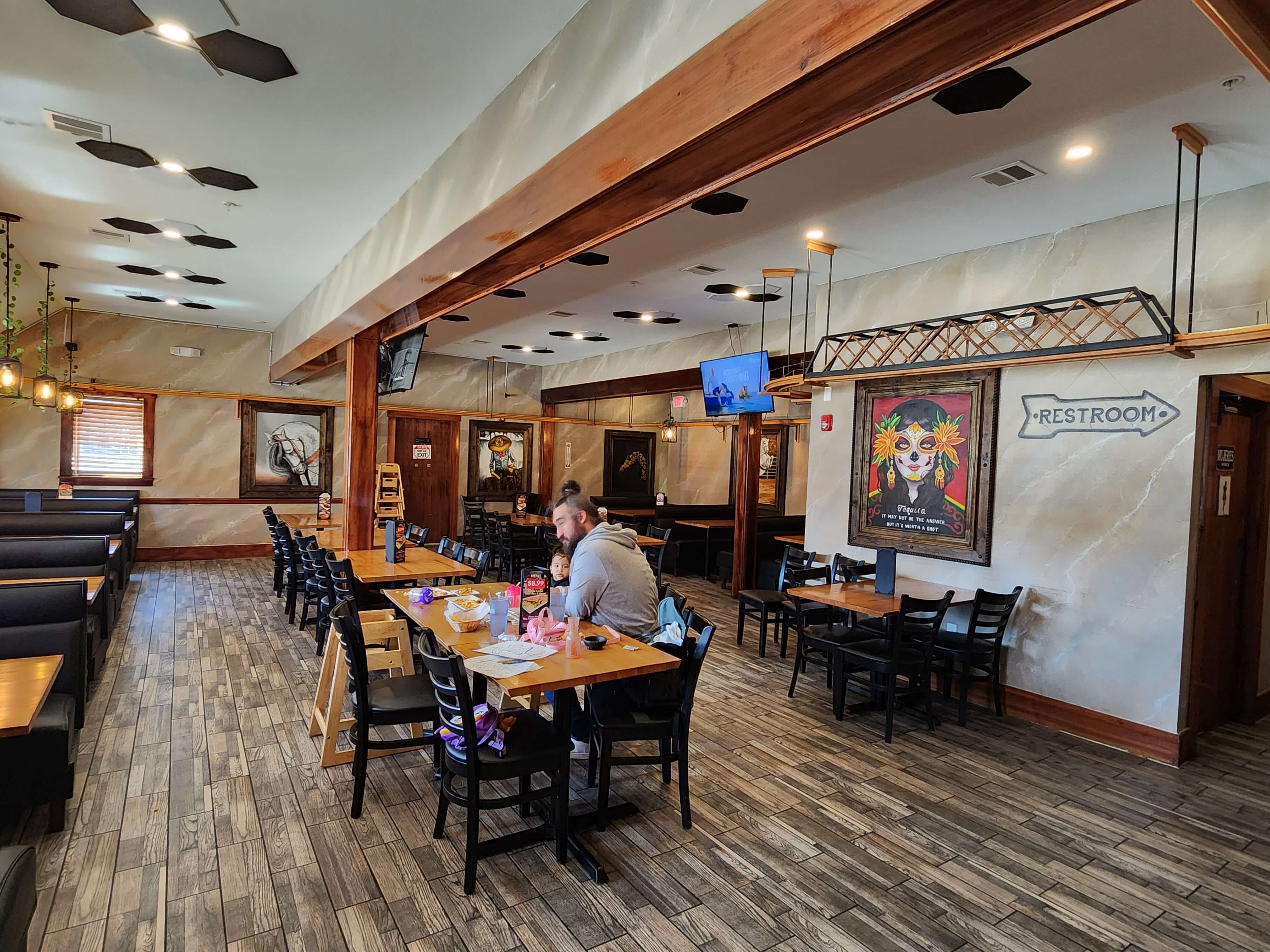 A spacious restaurant interior with wooden accents, featuring tables, booths, and a restroom sign, where a person is seated at a table.