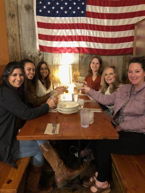 Six women smile and toast drinks at a table adorned with a rustic wooden backdrop and an American flag.