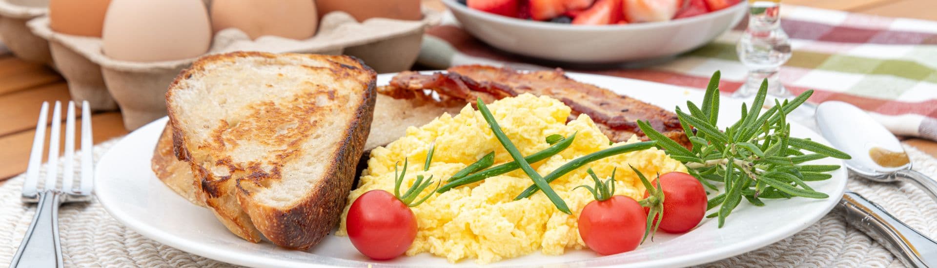 A plate of scrambled eggs, crispy bacon, toast, and cherry tomatoes, accompanied by a bowl of fresh fruit and a carton of eggs.