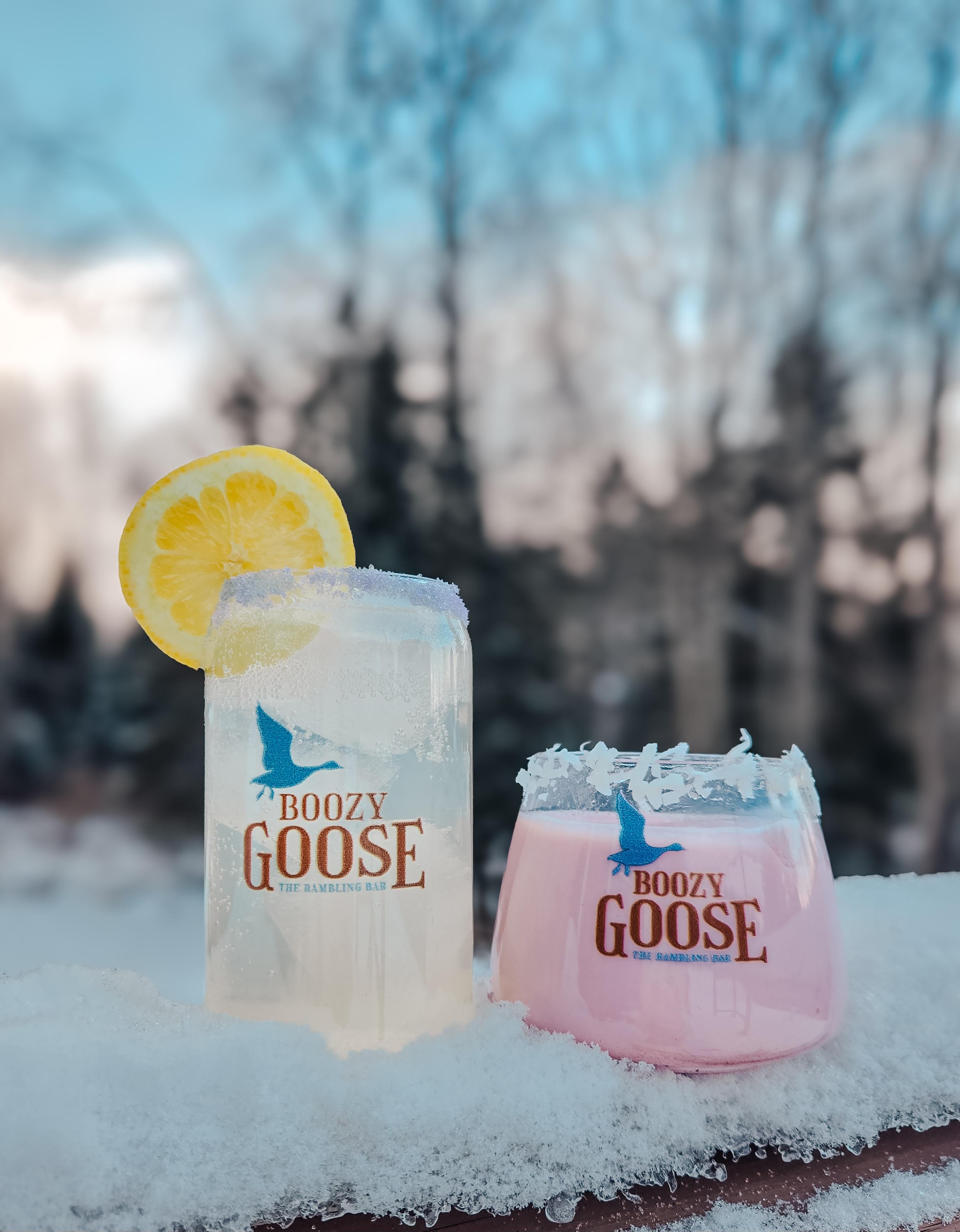 Two colorful cocktails sit on a snowy surface, one adorned with a lemon slice, both featuring the "Boozy Goose" logo.