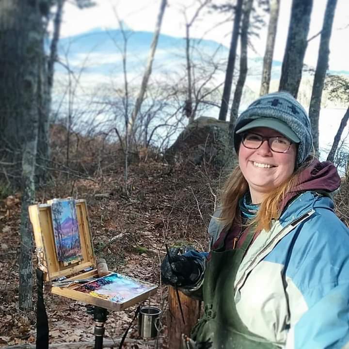 A smiling artist stands in a forest, holding a paintbrush beside an easel with a colorful landscape painting.