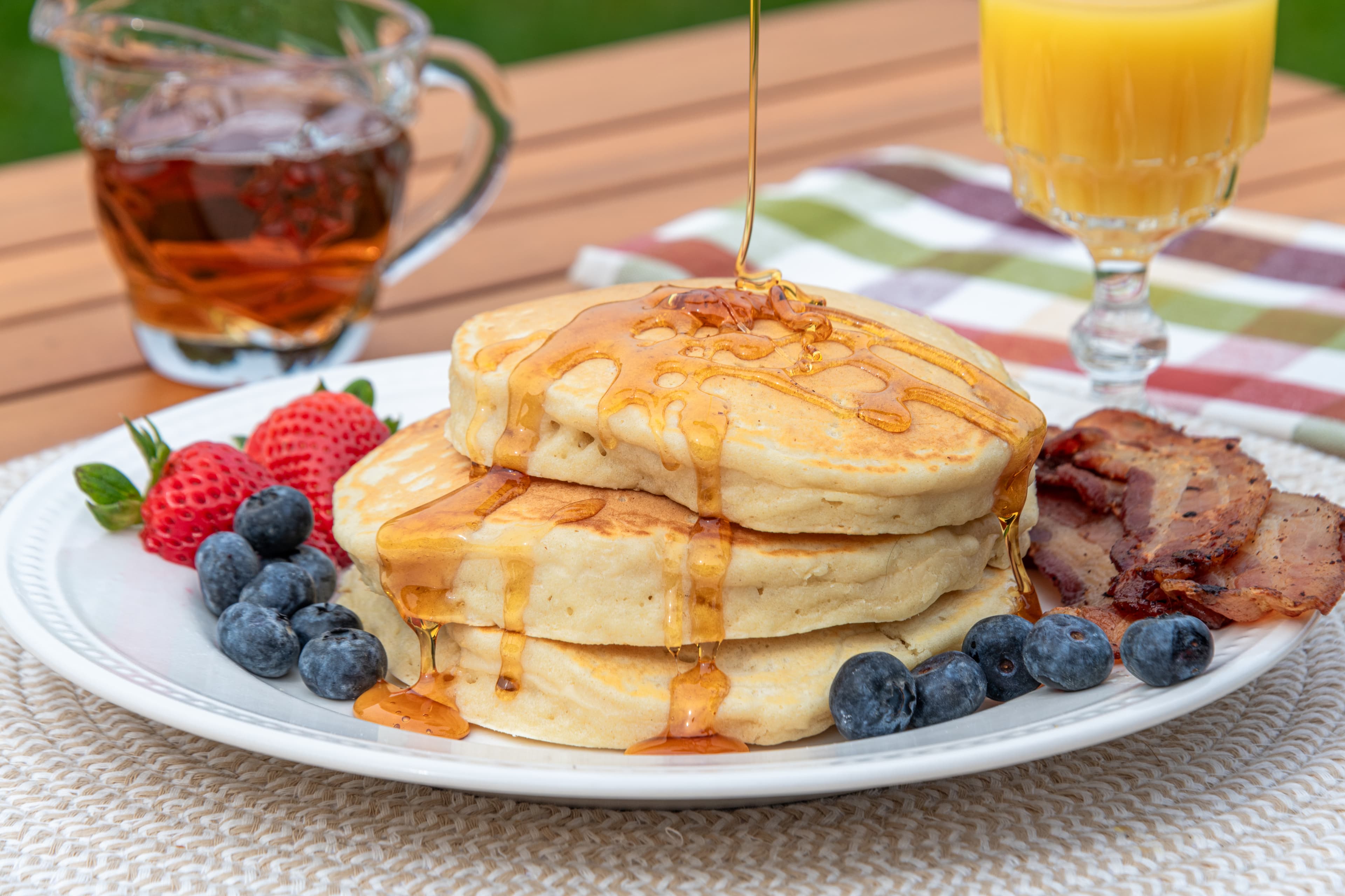 Stack of three fluffy, golden pancakes with blueberries, strawberries, bacon and a glass of orange juice.