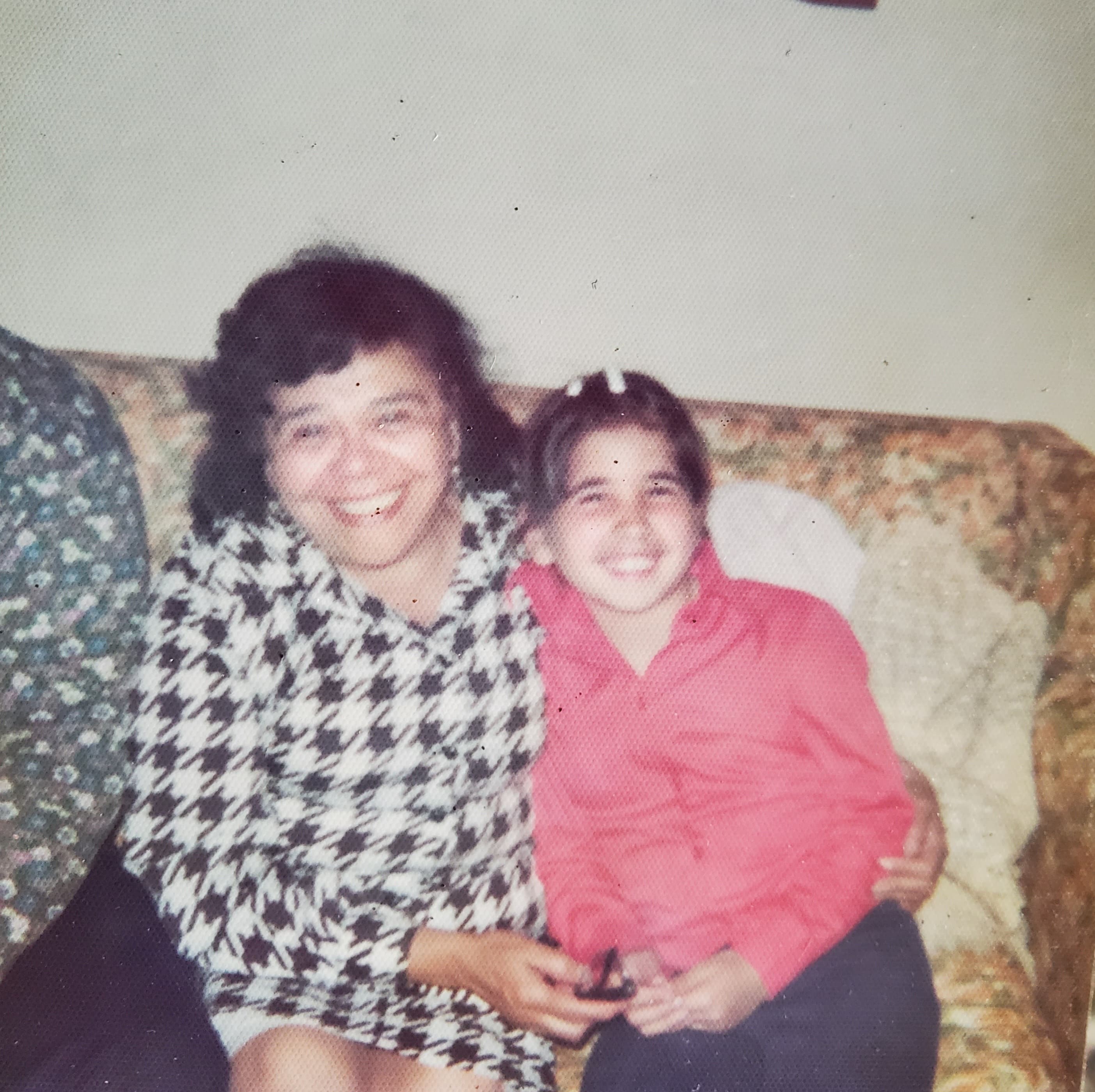 A smiling woman and girl sit closely together on a couch, sharing a joyful moment.