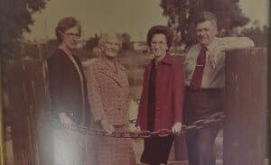 Four people stand together by a chain across a wooden fence, dressed in formal attire against a vintage backdrop.